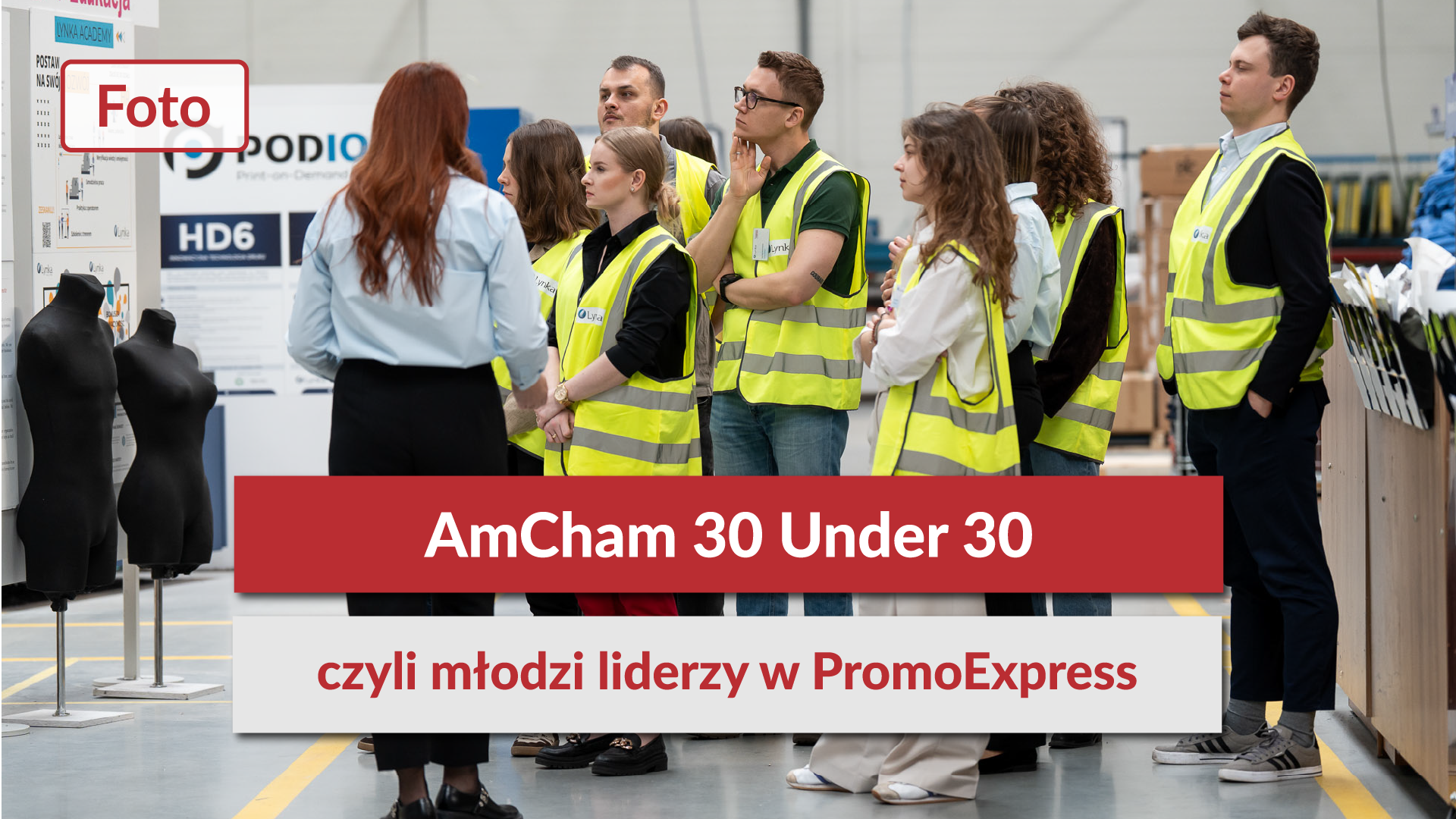 Zdjęcie główne z czerwonym polem tekstowym: „AmCham 30 Under 30 czyli młodzi liderzy w PromoExpress”. W tle grupa uczestników w kamizelkach odblaskowych zwiedzająca nowoczesną halę produkcyjną.