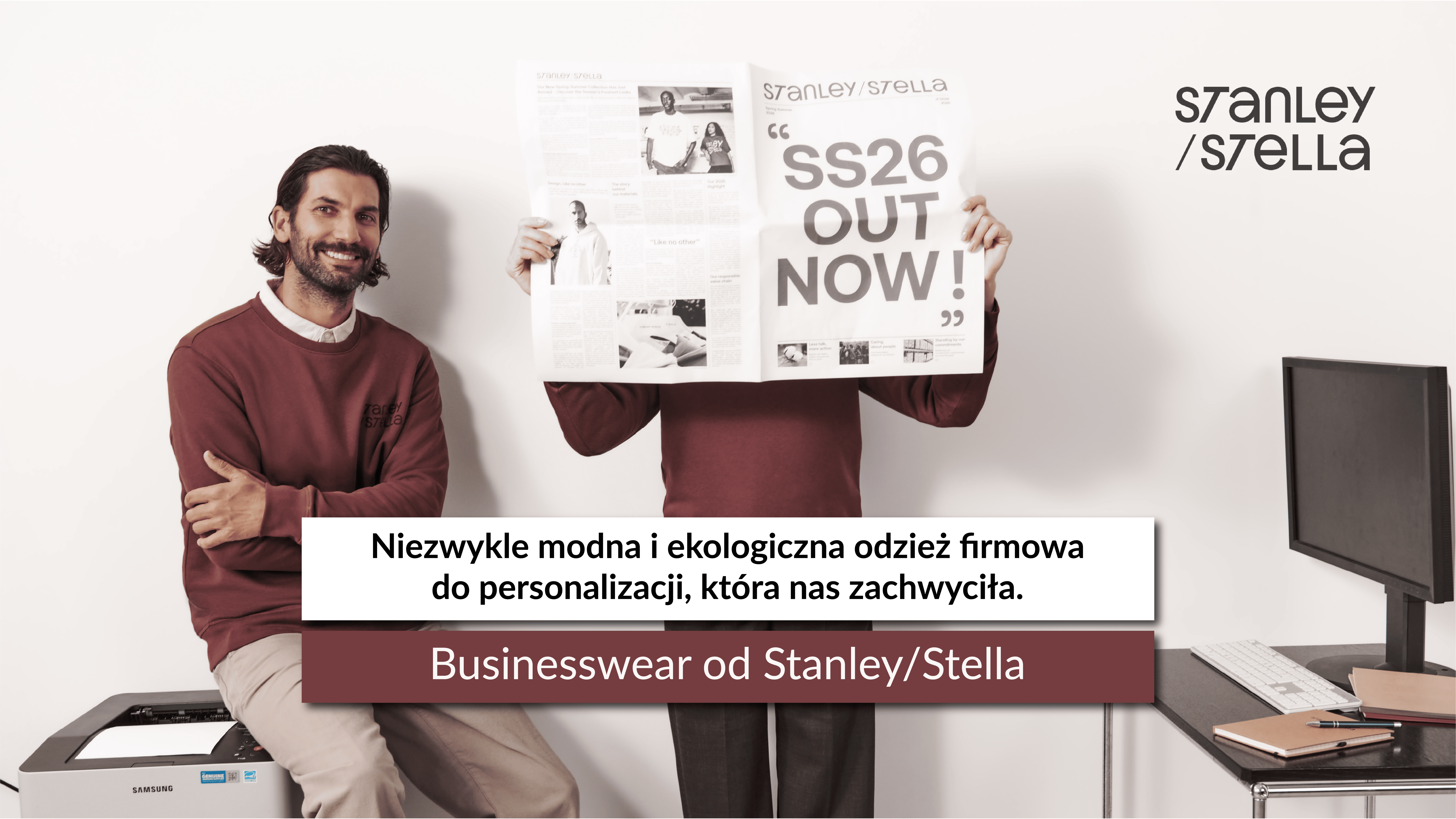 Mężczyzna w bordowej bluzie siedzi na drukarce obok osoby trzymającej gazetę z nagłówkiem „SS26 OUT NOW!”. Na środku napis dotyczący ekologicznej odzieży firmowej.