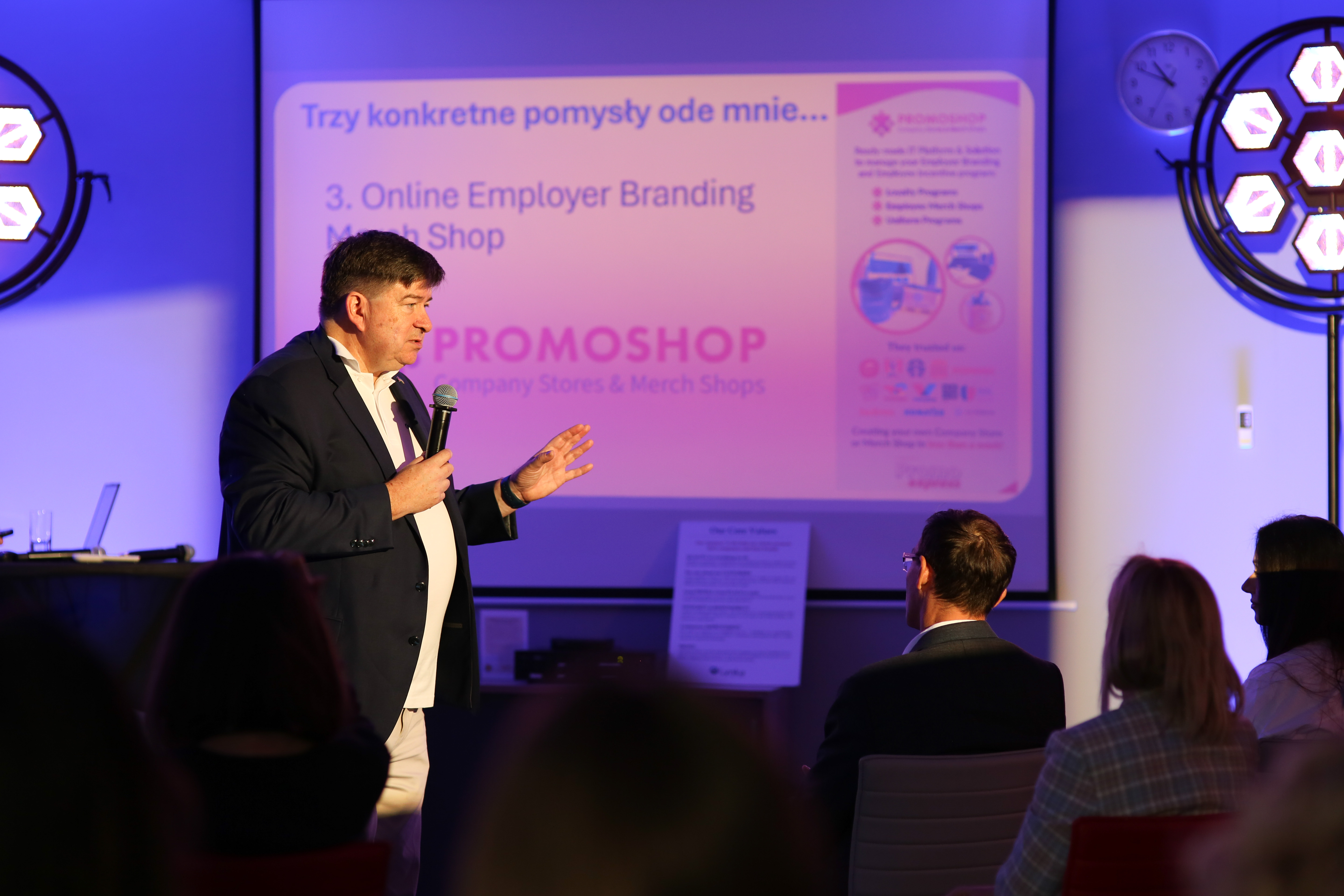 Katarzyna Luther podczas konferencji o Employer Branding, prezentacja na scenie z mikrofonem, w tle ekspozycja koszulek i kurtek.