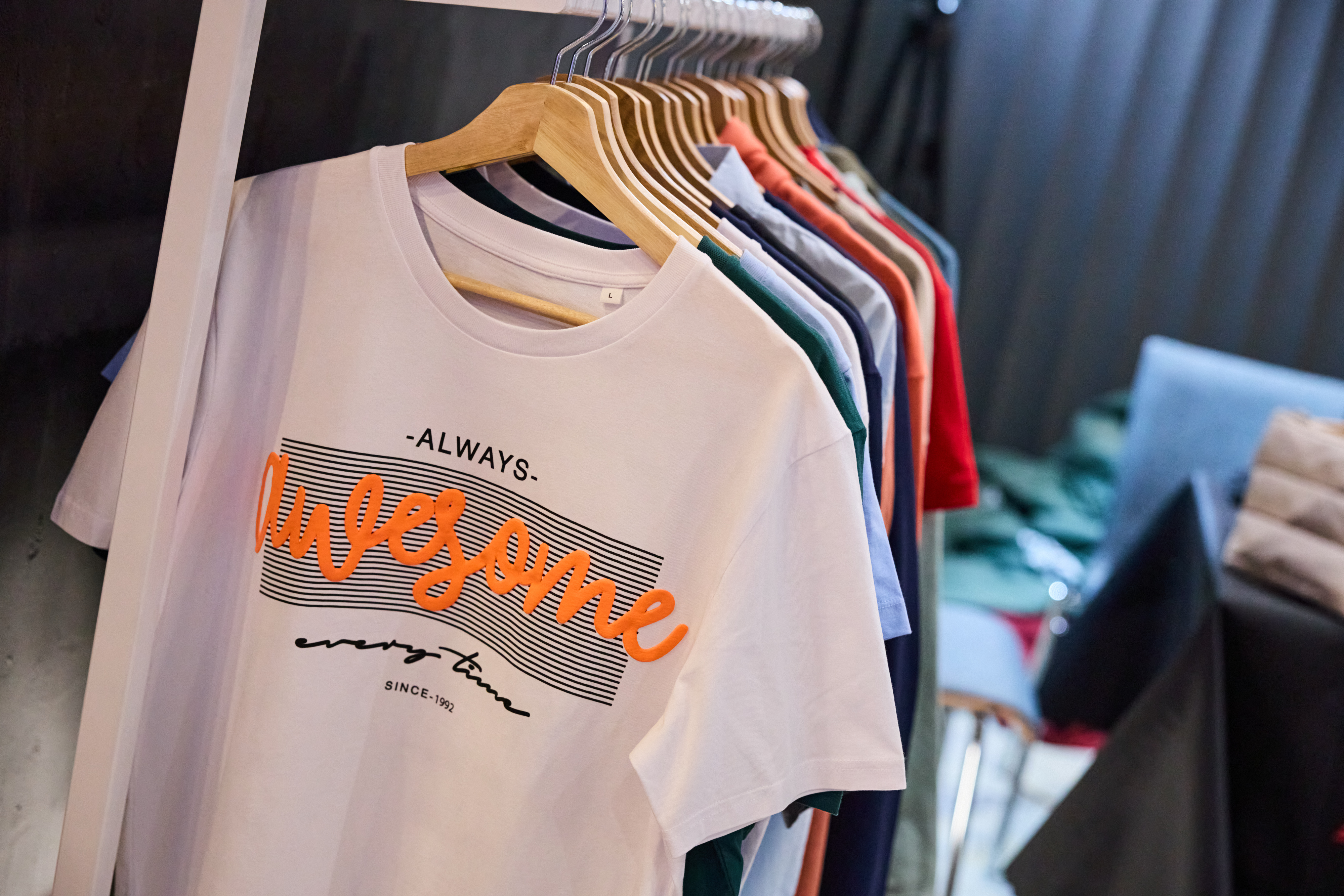 Zbliżenie na biały t-shirt z pomarańczowym wypukłym napisem &bdquo;Awesome&rdquo; na wieszaku. W tle widoczne inne kolorowe ubrania na stojaku w nowoczesnym wnętrzu.