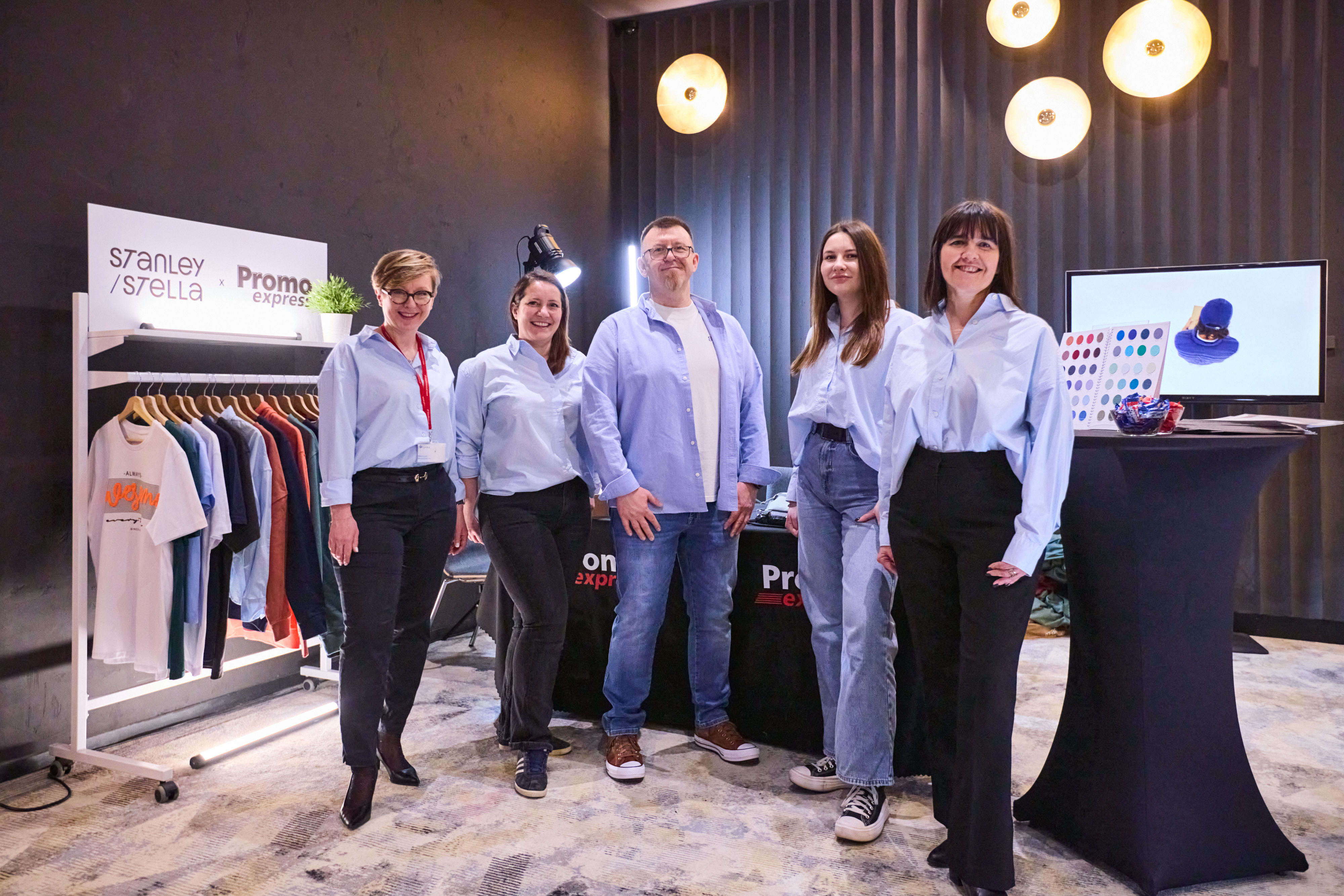 Pięcioosobowy zesp&oacute;ł Promo Express pozuje przy stoisku Stanley/Stella. Pracownicy są ubrani w błękitne koszule i jeansy lub ciemne spodnie. Obok stoi wieszak z odzieżą.