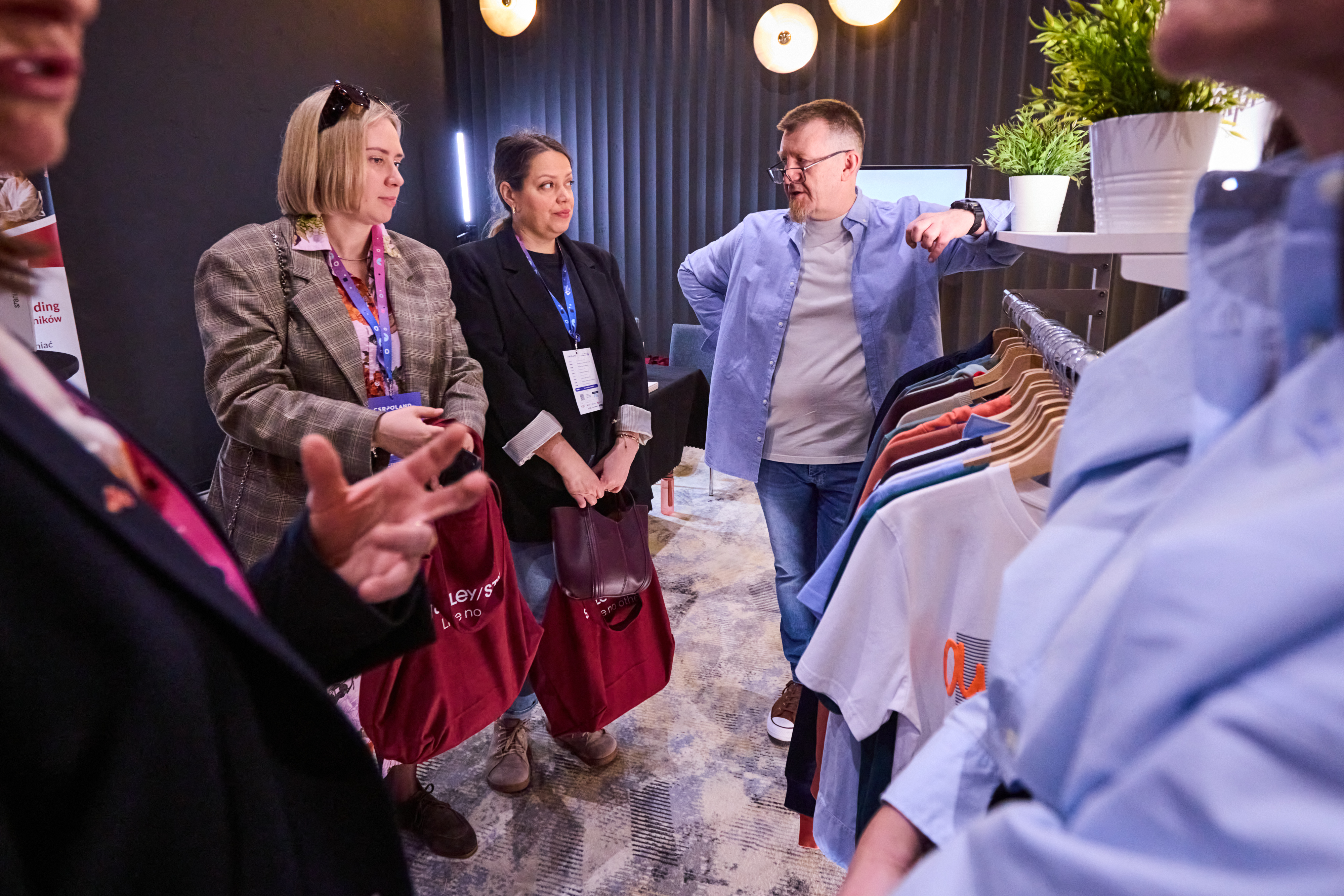 Mężczyzna z obsługi stoiska rozmawia z dwiema kobietami trzymającymi bordowe torby reklamowe. W tle widoczny wieszak z kolorowymi koszulkami i nowoczesne oświetlenie.