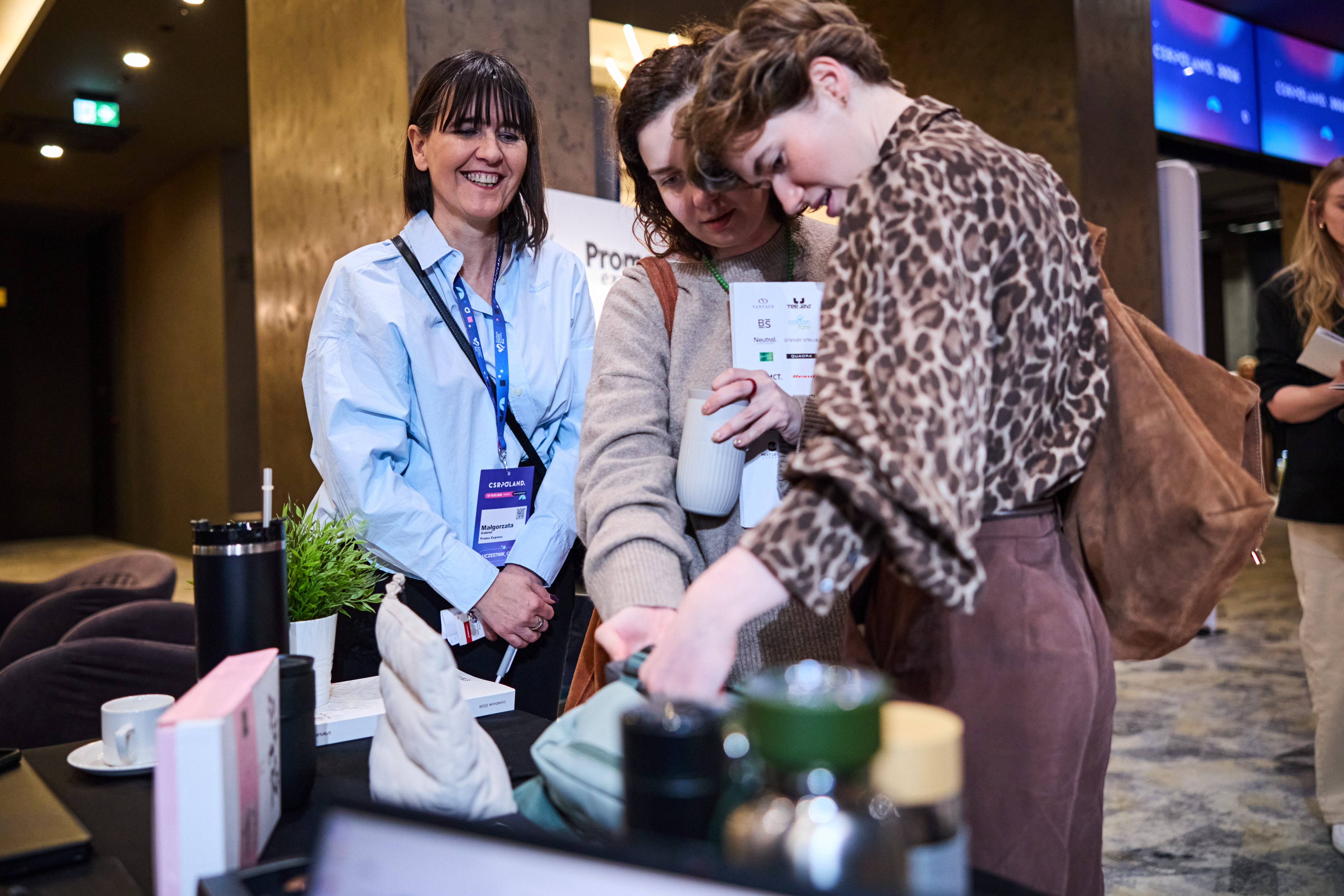 Uśmiechnięta kobieta z identyfikatorem prezentuje ofertę stoiska dw&oacute;m klientkom. Na pierwszym planie widoczne akcesoria konferencyjne, kubek i rośliny doniczkowe.
