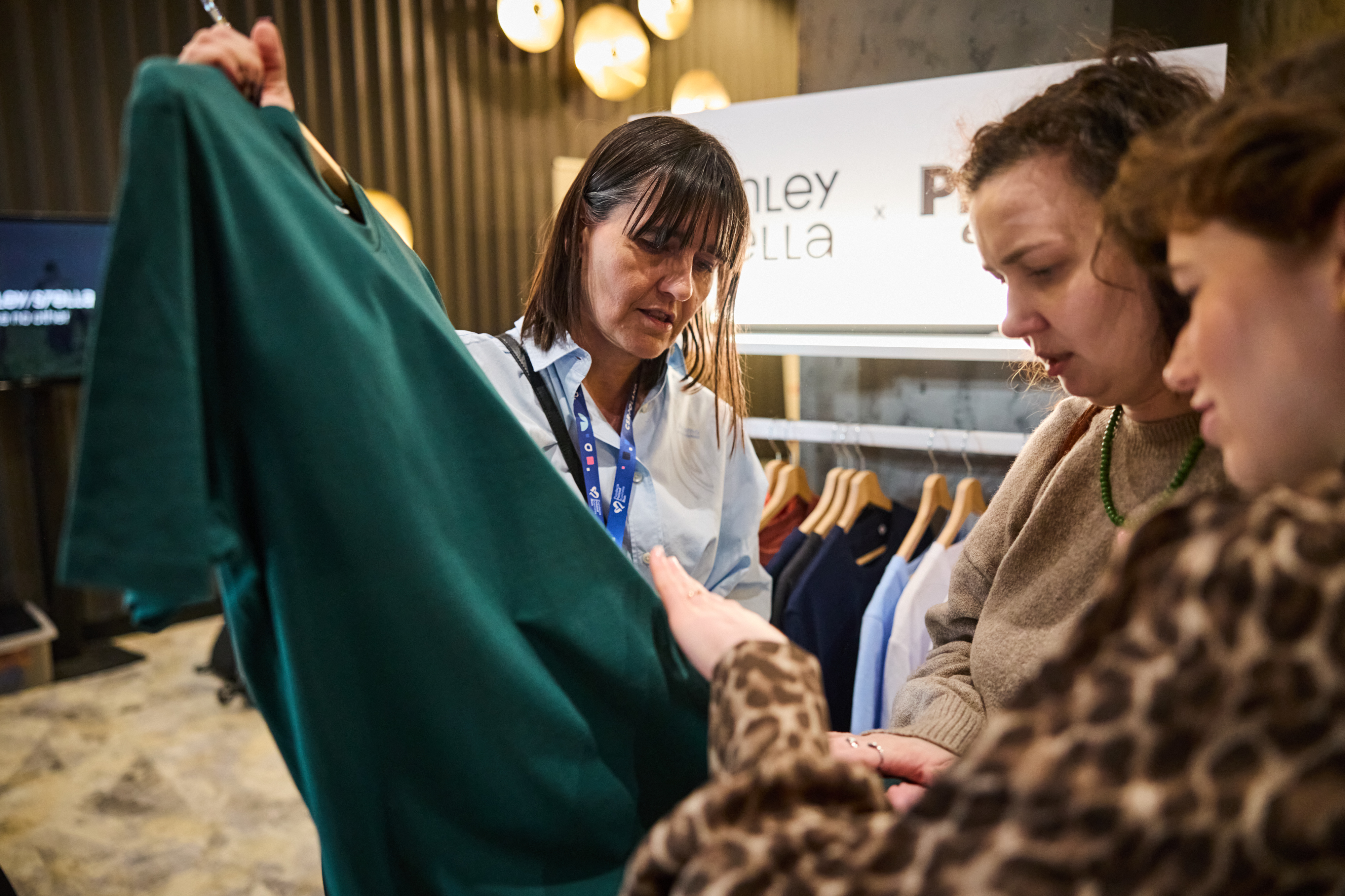 Przedstawicielka marki prezentuje klientkom ciemnozieloną koszulkę zdjętą z wieszaka. Kobiety z uwagą oglądają materiał i jakość wykonania produktu na stoisku.