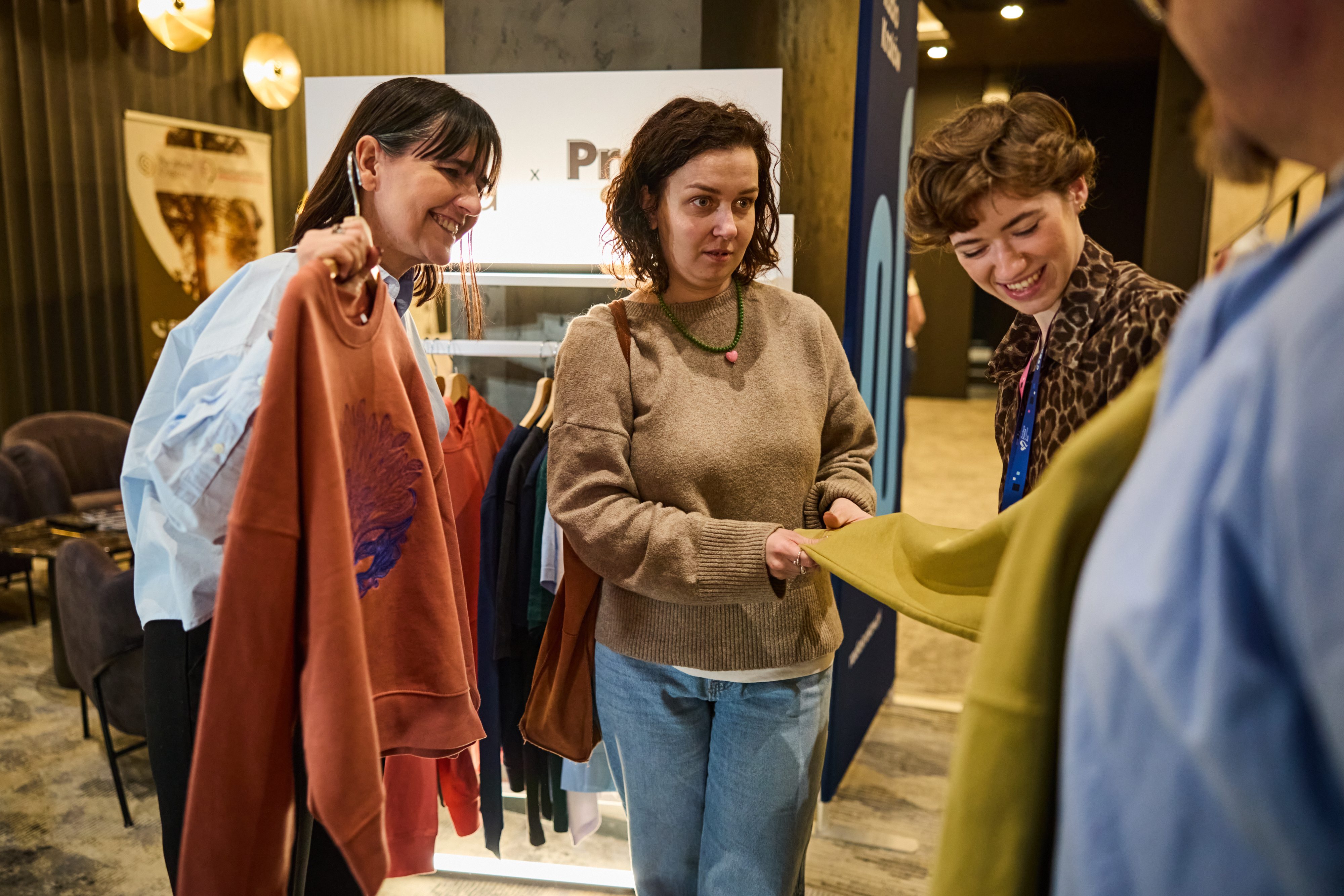 Trzy kobiety oglądają i dotykają kolorowe bluzy na stoisku Promo Express. Jedna z nich trzyma rdzawą bluzę z nadrukiem, prezentując ją pozostałym osobom.