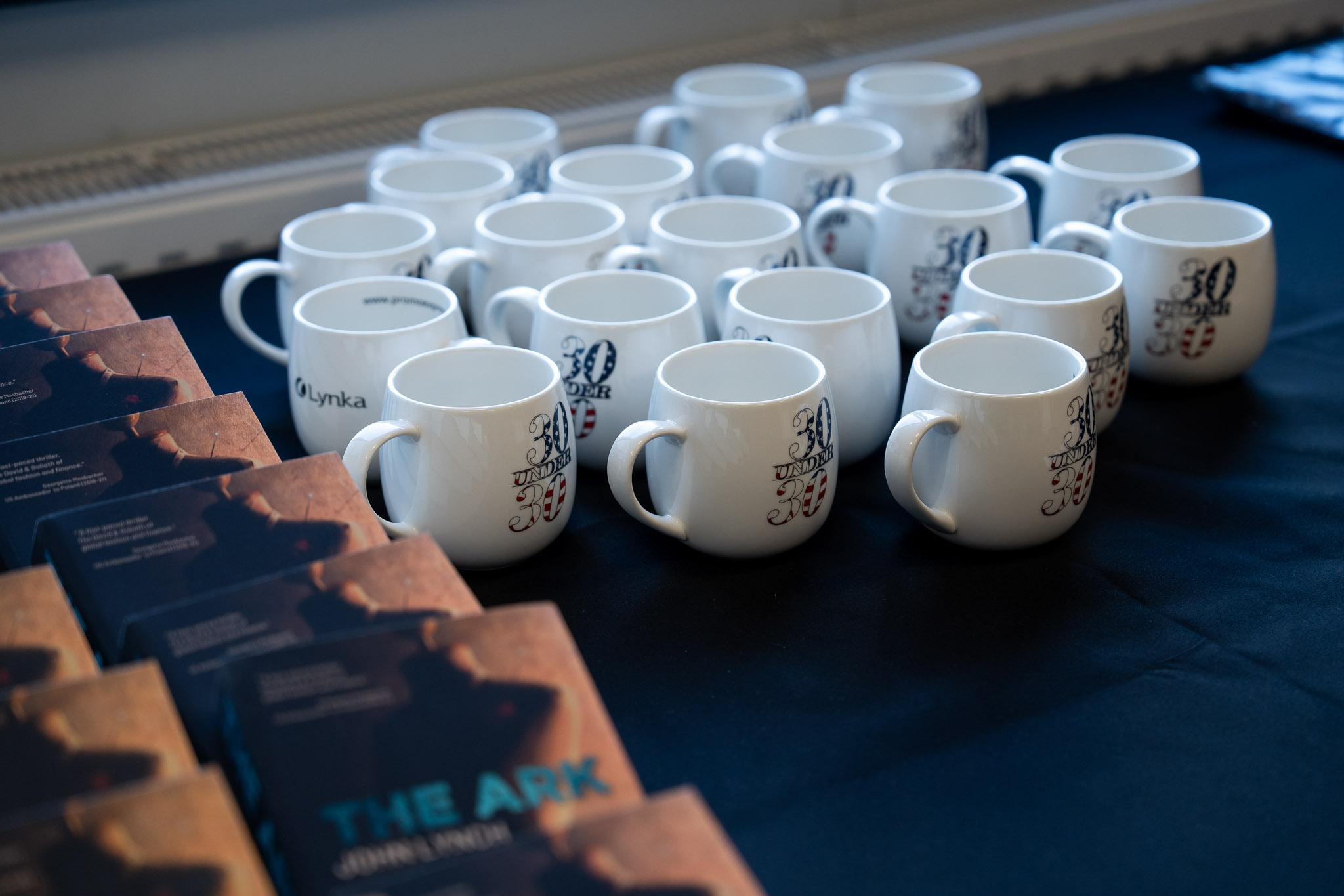 Białe filiżanki z logo AmCham ułożone w rzędach na czarnym obrusie.