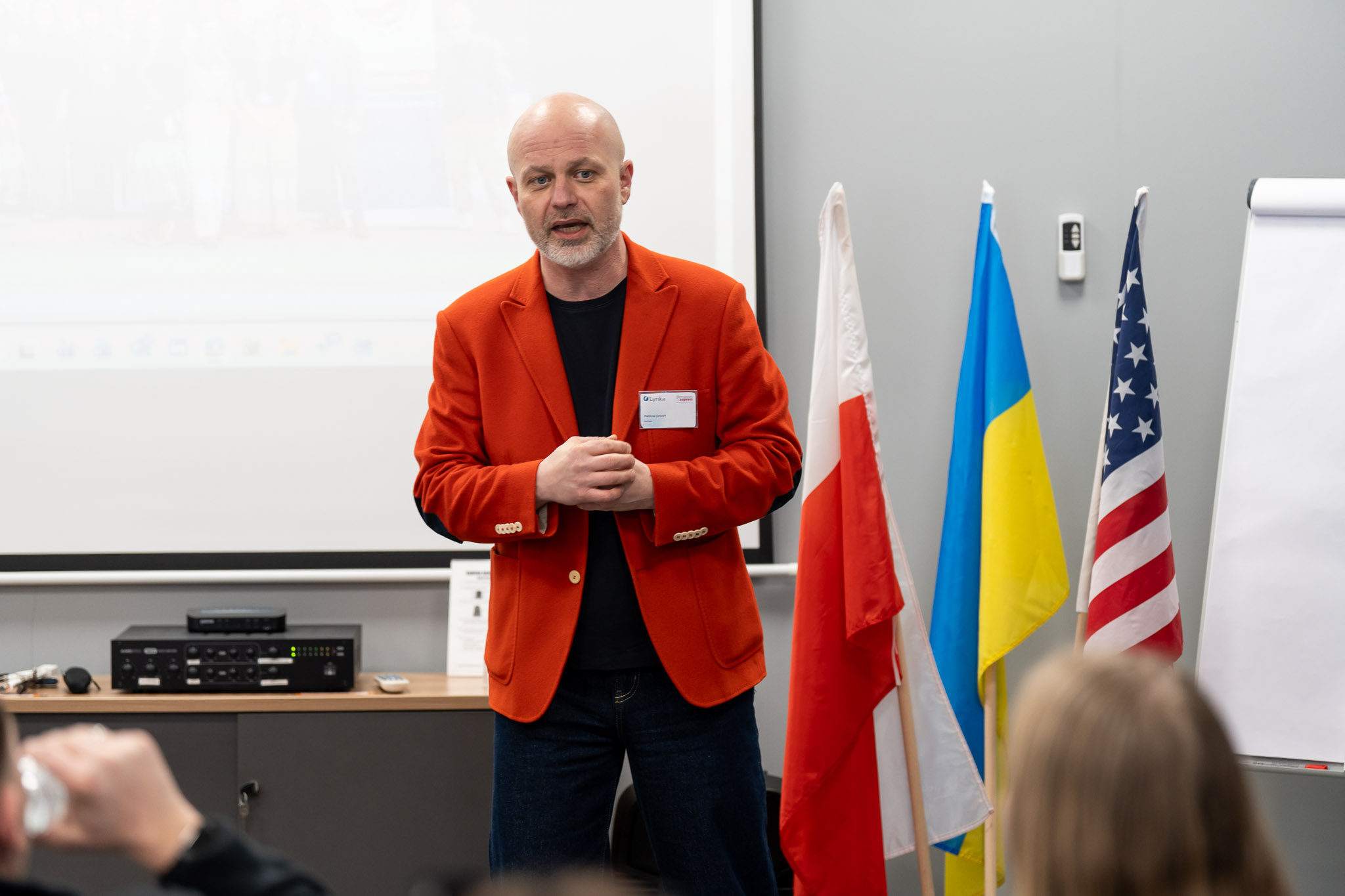 Prelegent w czerwonej marynarce przemawiający na tle flag Polski, Ukrainy i USA.