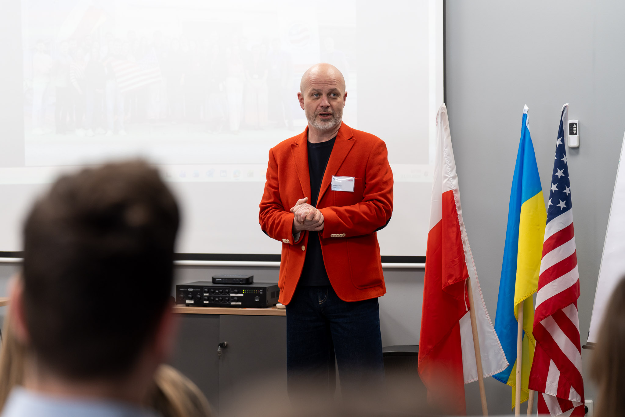Wystąpienie otwierające w sali konferencyjnej, prelegent w czerwonej marynarce.
