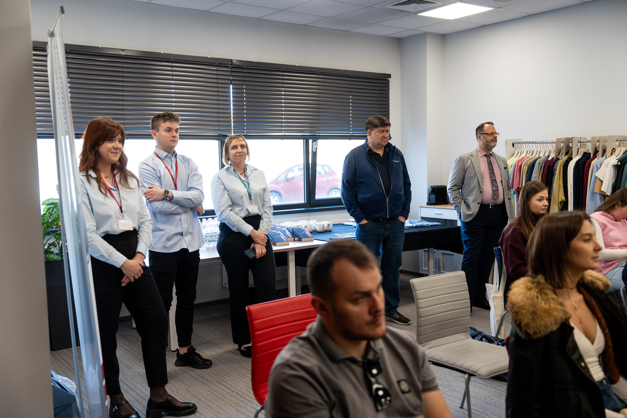 Grupa uczestnik&oacute;w stojąca wzdłuż ściany podczas prezentacji.