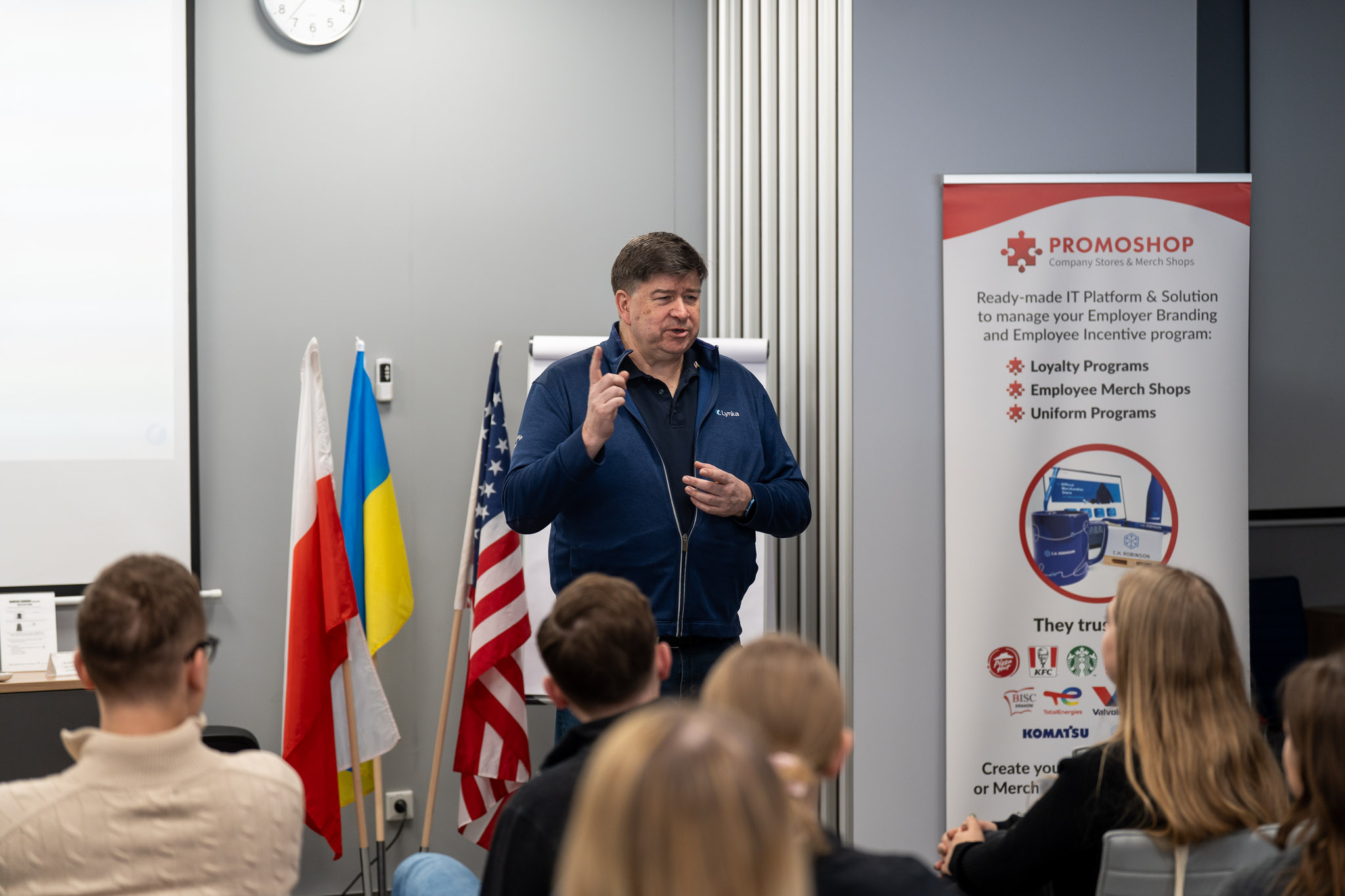 Prelegent przemawiający obok baneru AmCham i flag narodowych.