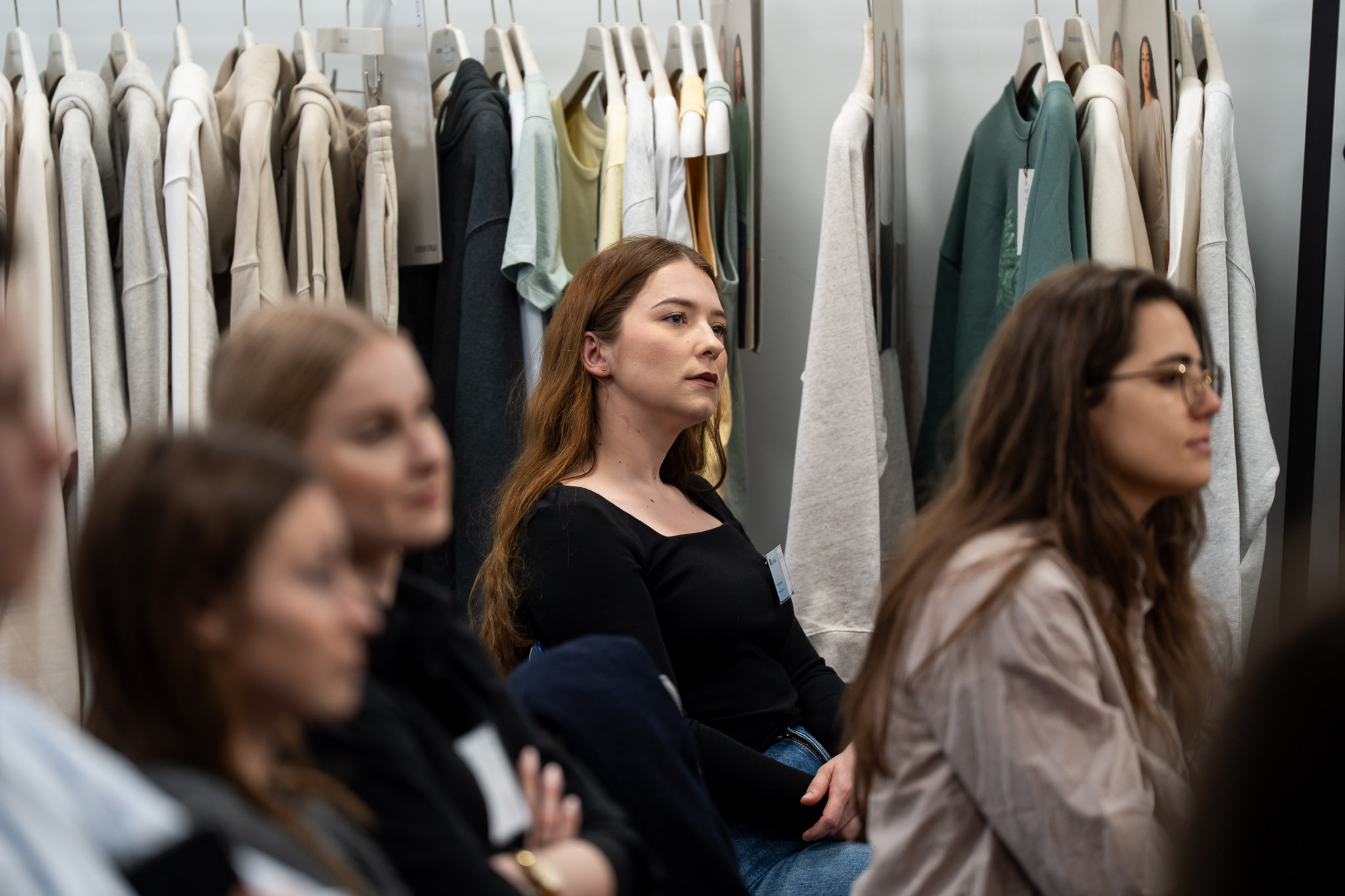 Trzy uczestniczki słuchające prelegenta na tle wieszak&oacute;w z pr&oacute;bkami odzieży.