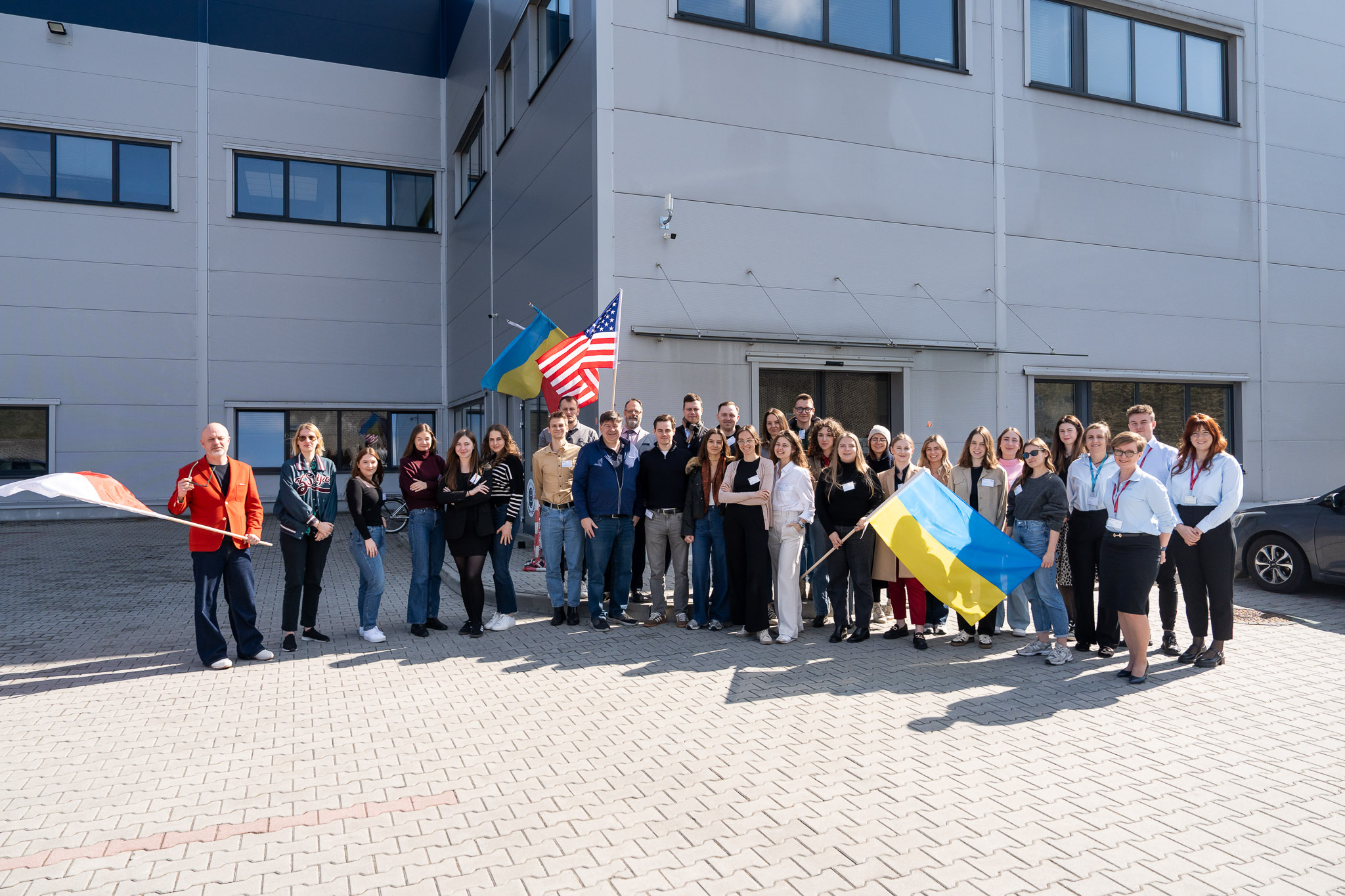 Duże zdjęcie grupowe wszystkich uczestnik&oacute;w przed budynkiem firmy z flagami.