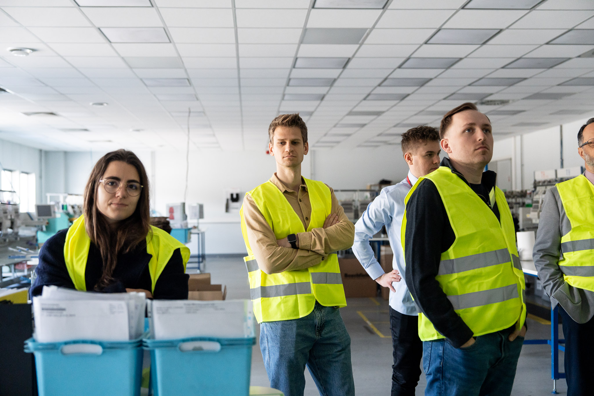 Młodzi uczestnicy w strojach ochronnych na tle regał&oacute;w magazynowych.