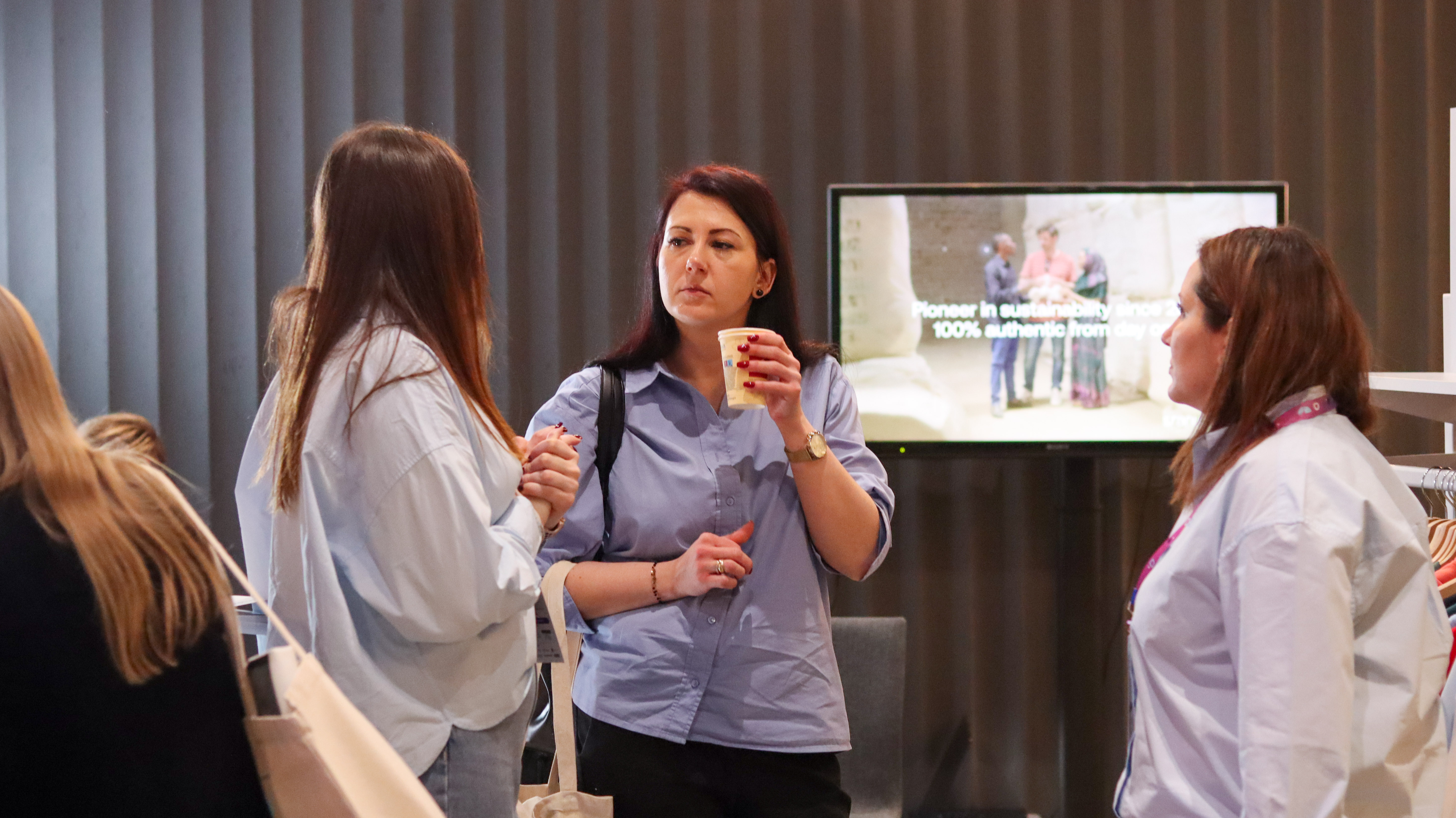 Trzy kobiety rozmawiają w strefie konferencyjnej. Jedna z nich pije kawę z papierowego kubka. W tle monitor wyświetlający prezentację o zr&oacute;wnoważonym rozwoju.