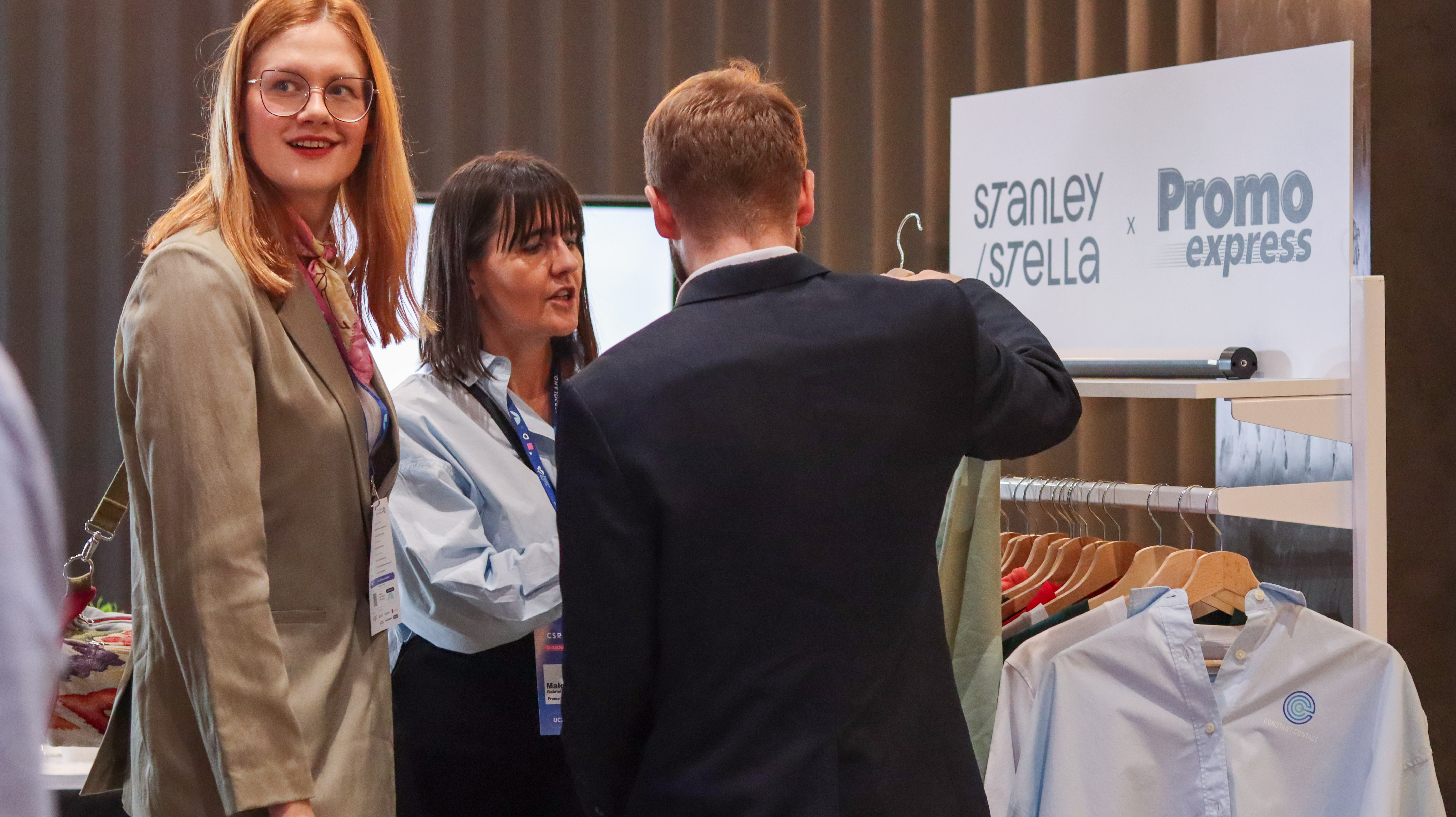Zbliżenie na stoisko z odzieżą reklamową. Szereg kolorowych bluz i koszulek marki Stanley/Stella wisi na białych wieszakach. Nad nimi biały baner &bdquo;STANLEY/STELLA x Promo express&rdquo;. Po lewej stronie widać osoby oglądającye ofertę.