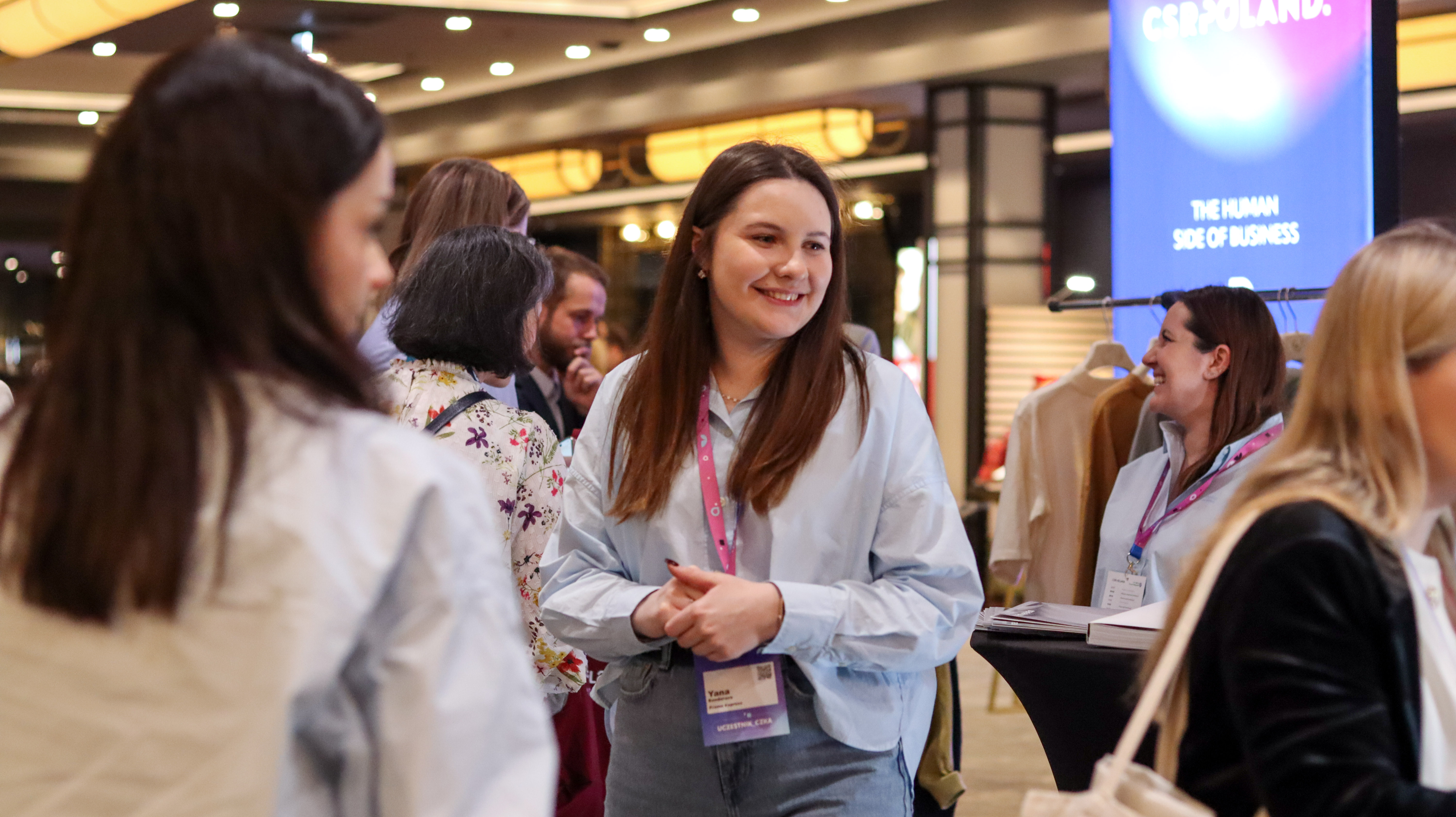 Grupa kobiet rozmawia w przestronnym lobby podczas wydarzenia CSR Poland. W tle widoczne nowoczesne oświetlenie sufitowe, inne stoiska oraz świecący totem reklamowy.