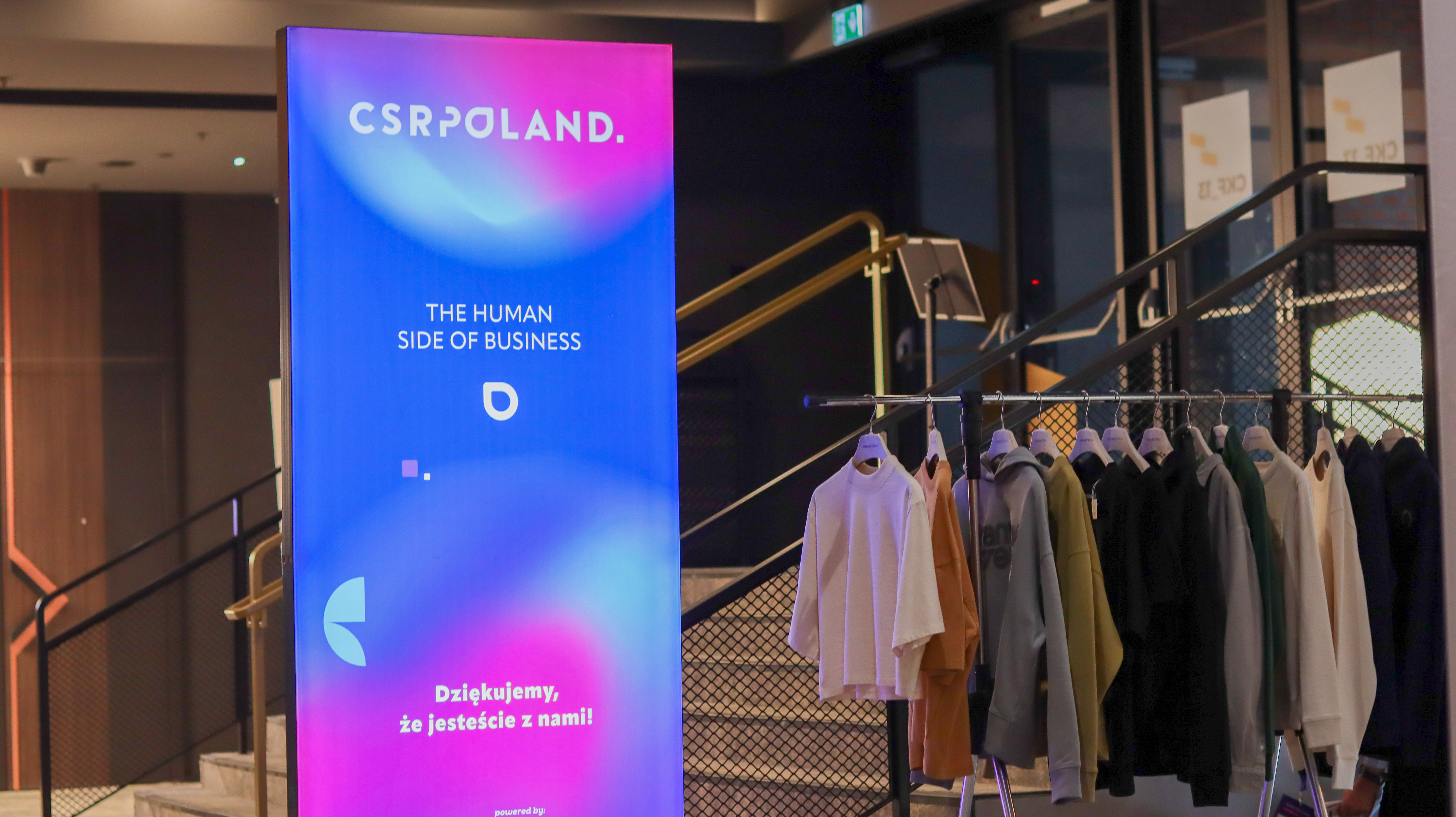 Świecący totem reklamowy CSR Poland z napisem "The Human Side of Business". W tle schody oraz wieszak z kolorową odzieżą marki Stanley/Stella.