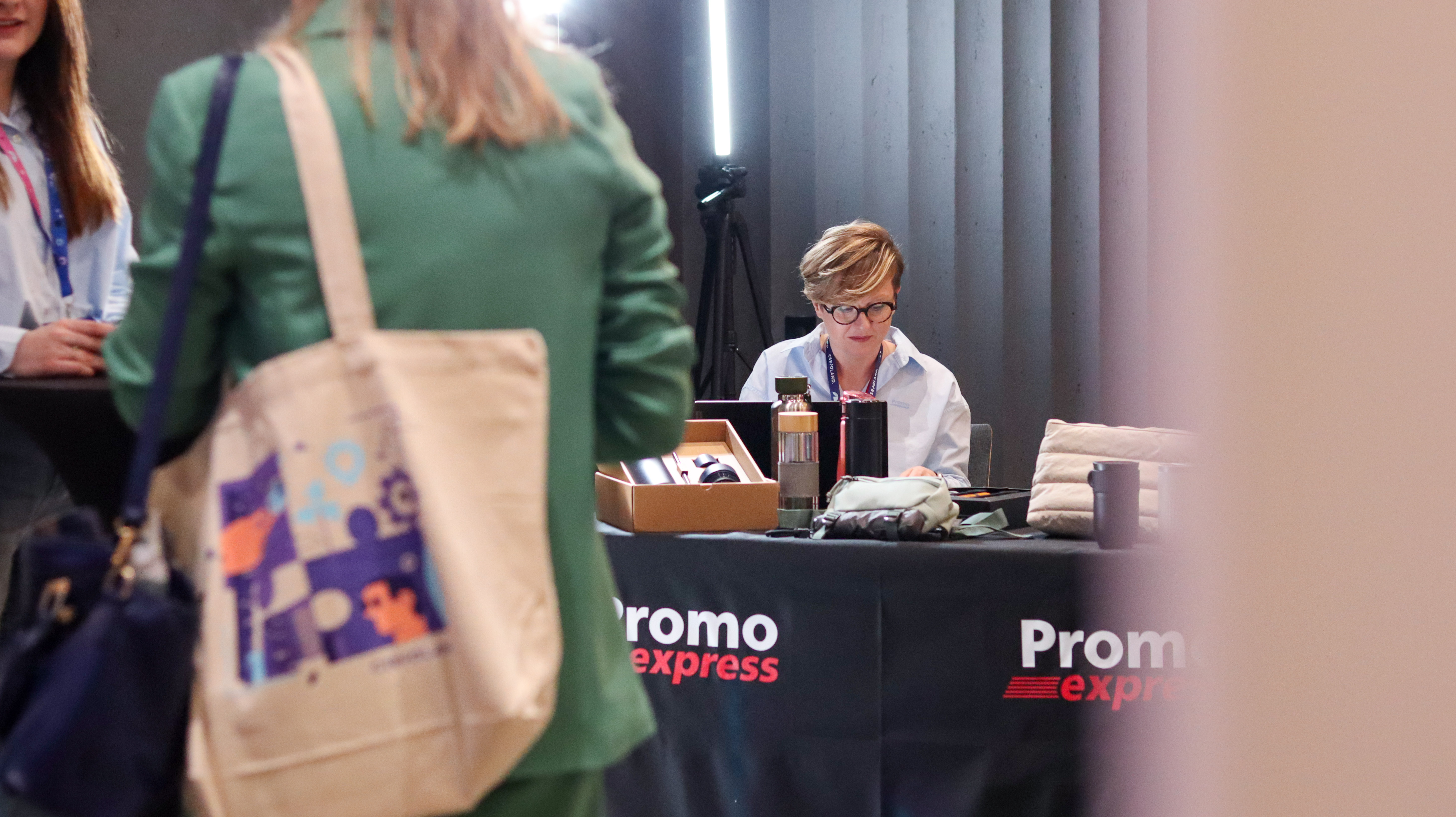 Zbliżenie na kobietę w okularach i błękitnej koszuli firmowej Promo Express podczas rozmowy z uczestnikiem konferencji.