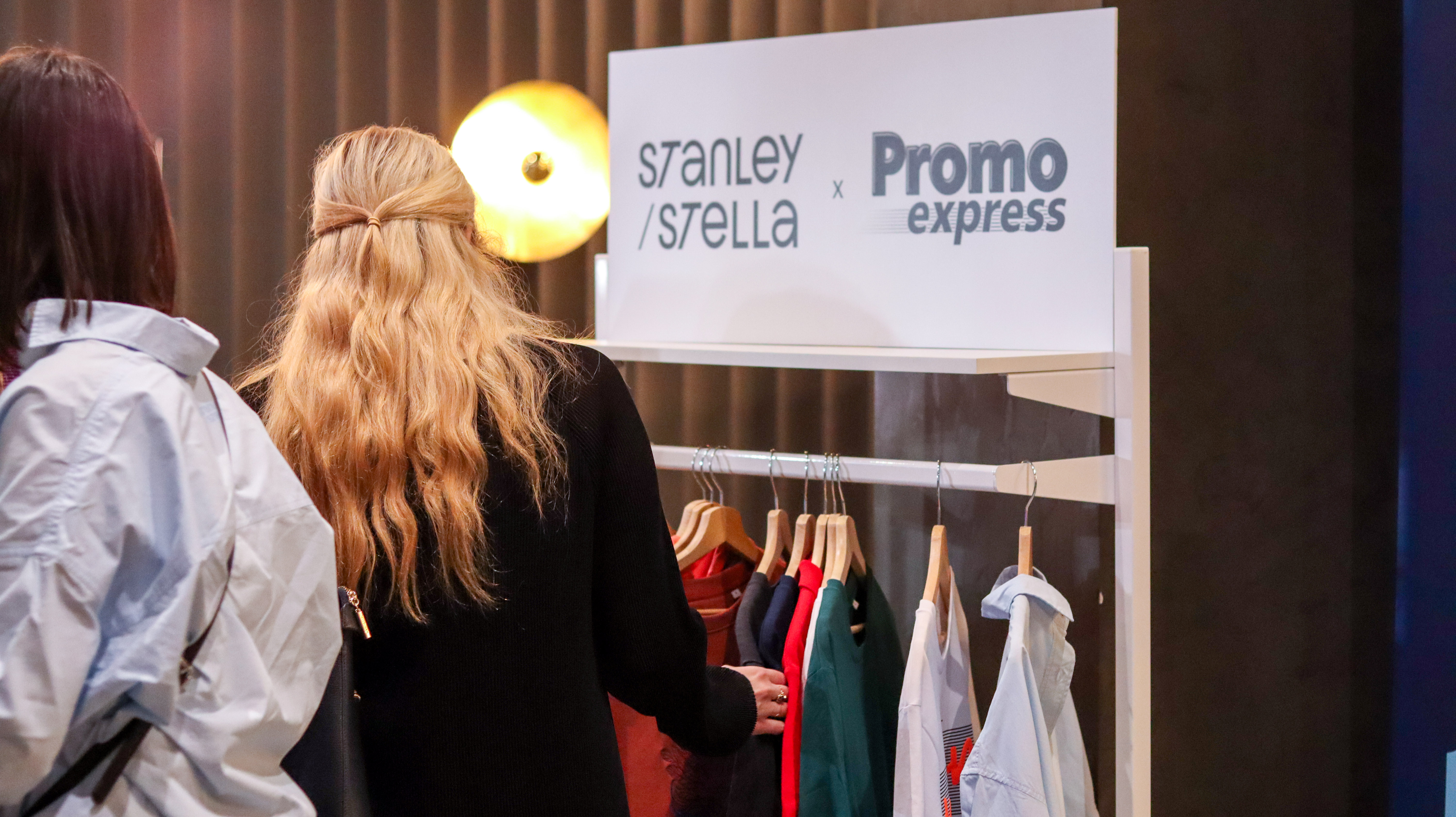 Blond kobieta na tle logotyp&oacute;w Stanley/Stella oraz Promo Express. Poniżej widoczny rząd kolorowych ubrań na wieszakach.