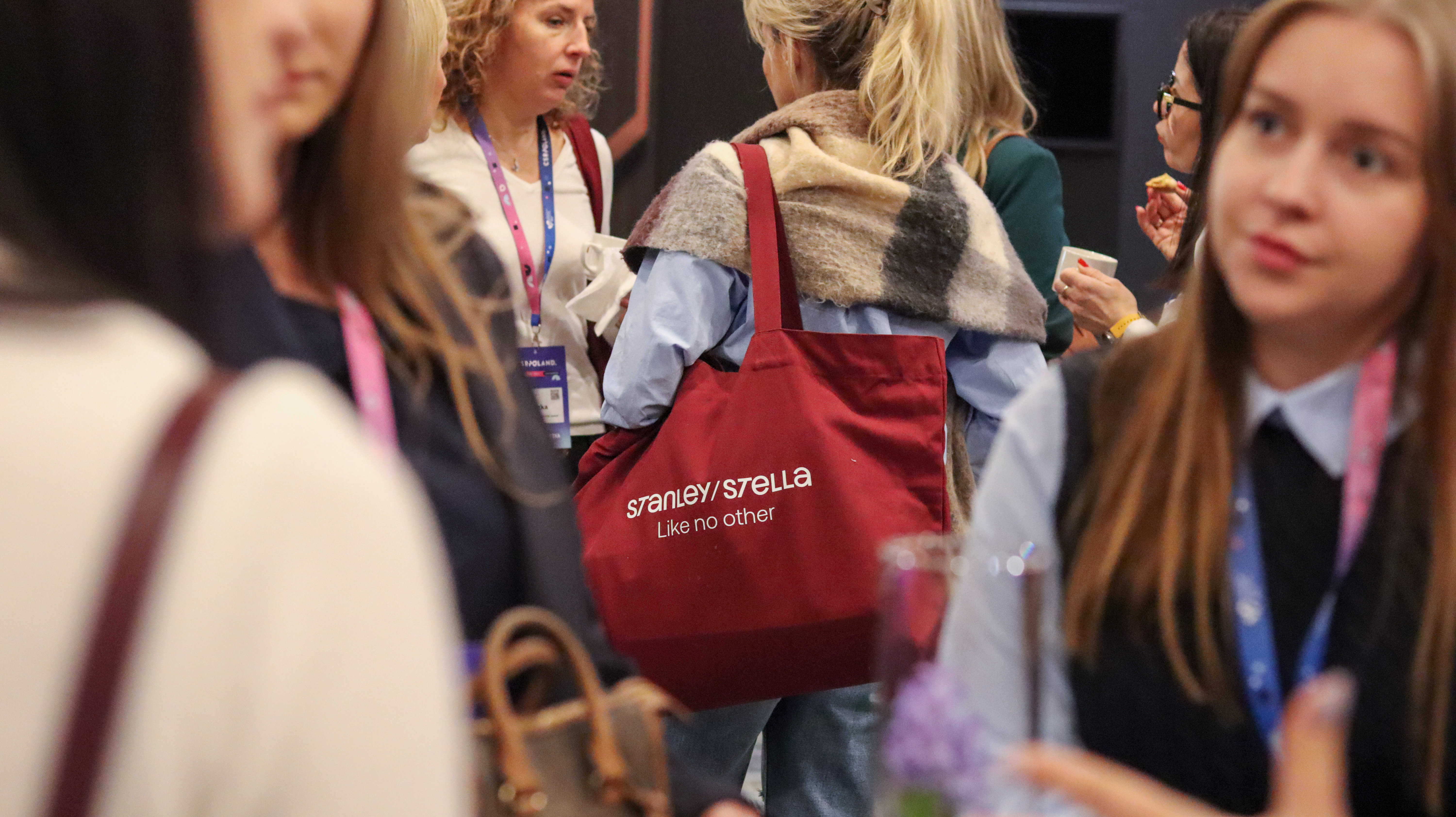 Uczestnicy wydarzenia w tłumie. Centralnie widoczna bordowa torba Promo Express na ramieniu kobiety w błękitnej koszuli. Na torbie biały nadruk &bdquo;STANLEY/STELLA Like no other&rdquo;. W tle widać rozmyte twarze i identyfikatory innych os&oacute;b.