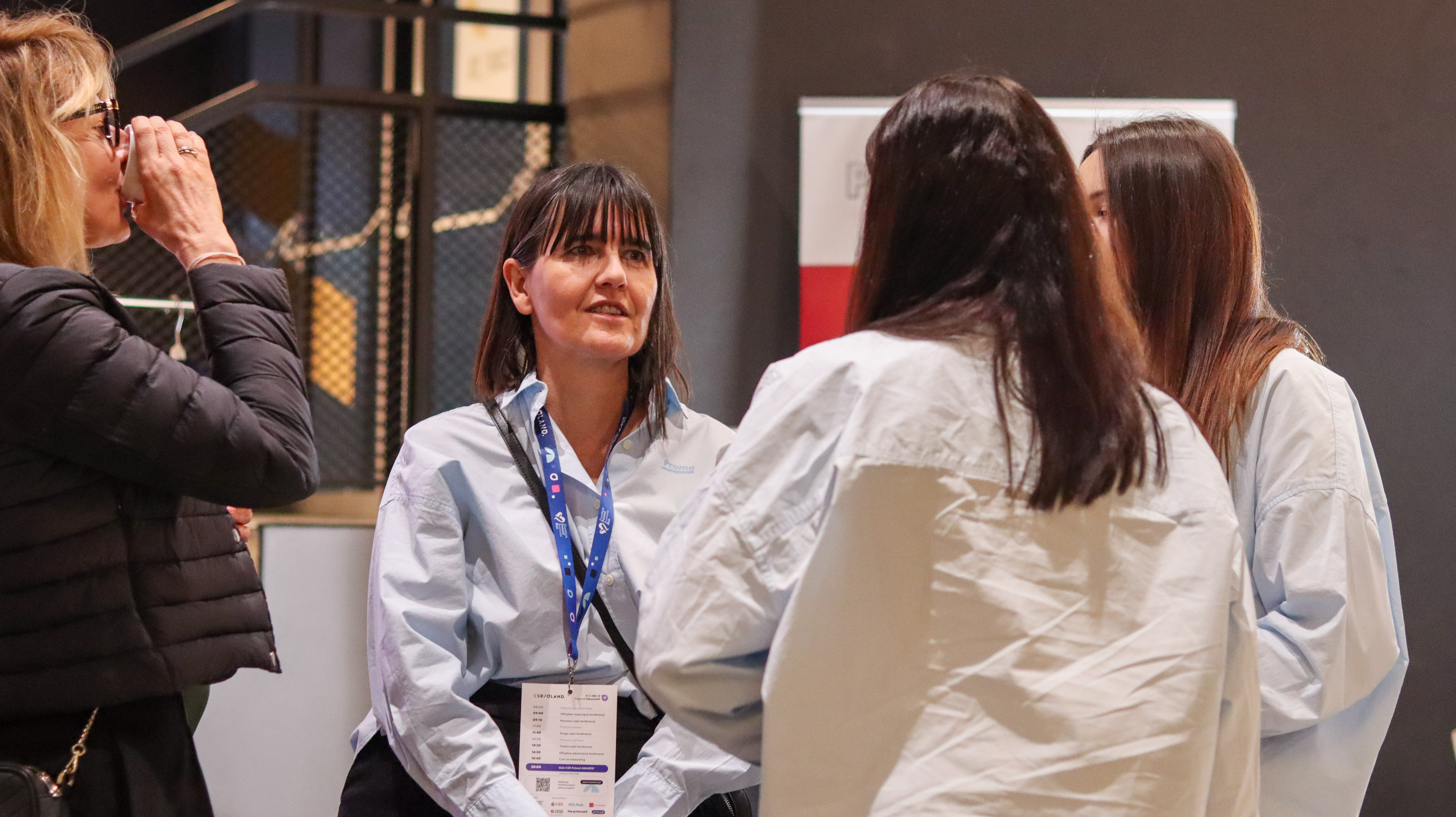 Kobieta w błękitnej koszuli z identyfikatorem rozmawia z grupą os&oacute;b podczas wydarzenia CSR Poland. W tle widoczne materiały promocyjne.