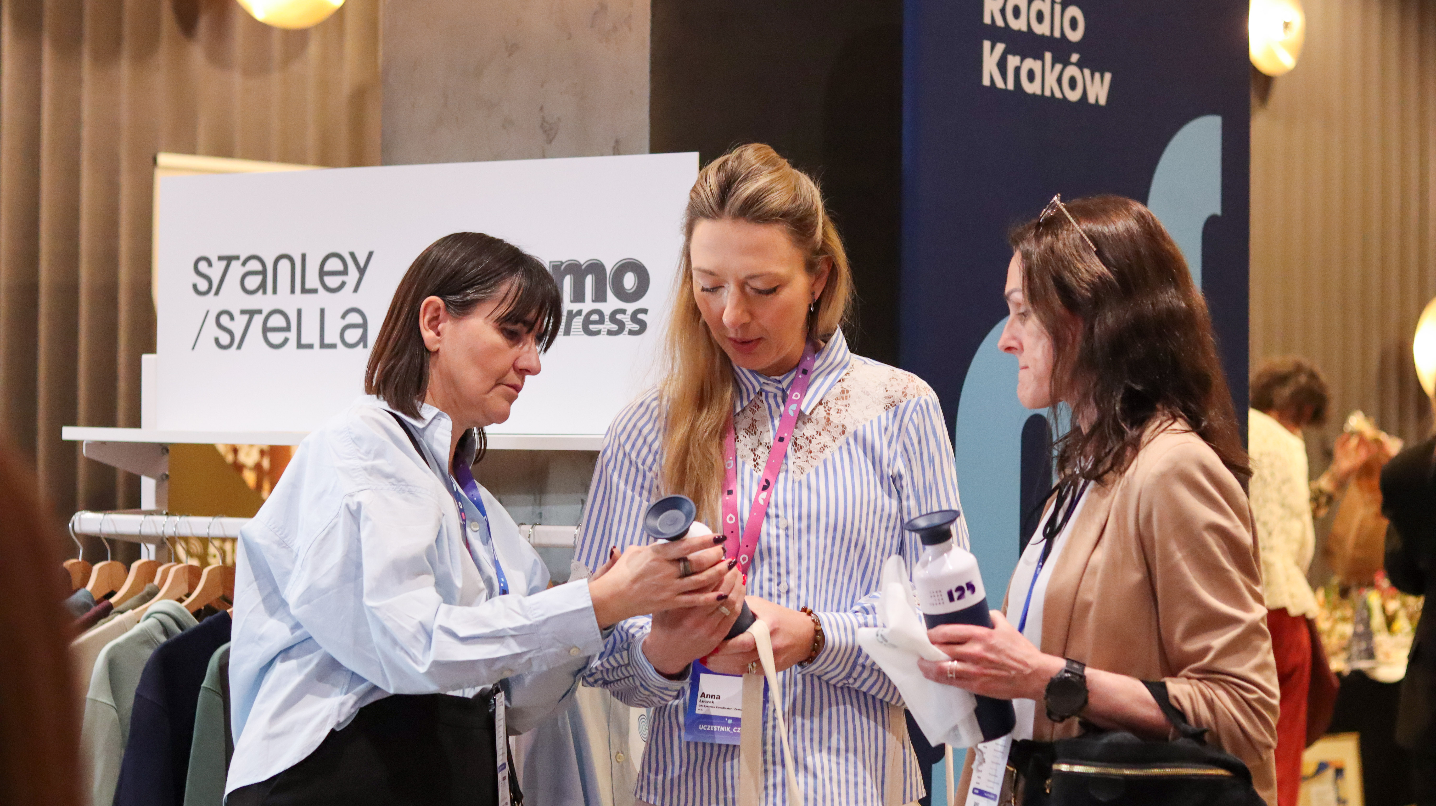 Rozmowa przy stoisku Promo Express. Przedstawicielka firmy w błękitnej koszuli prezentuje ofertę gadżet&oacute;w uczestniczce wydarzenia.