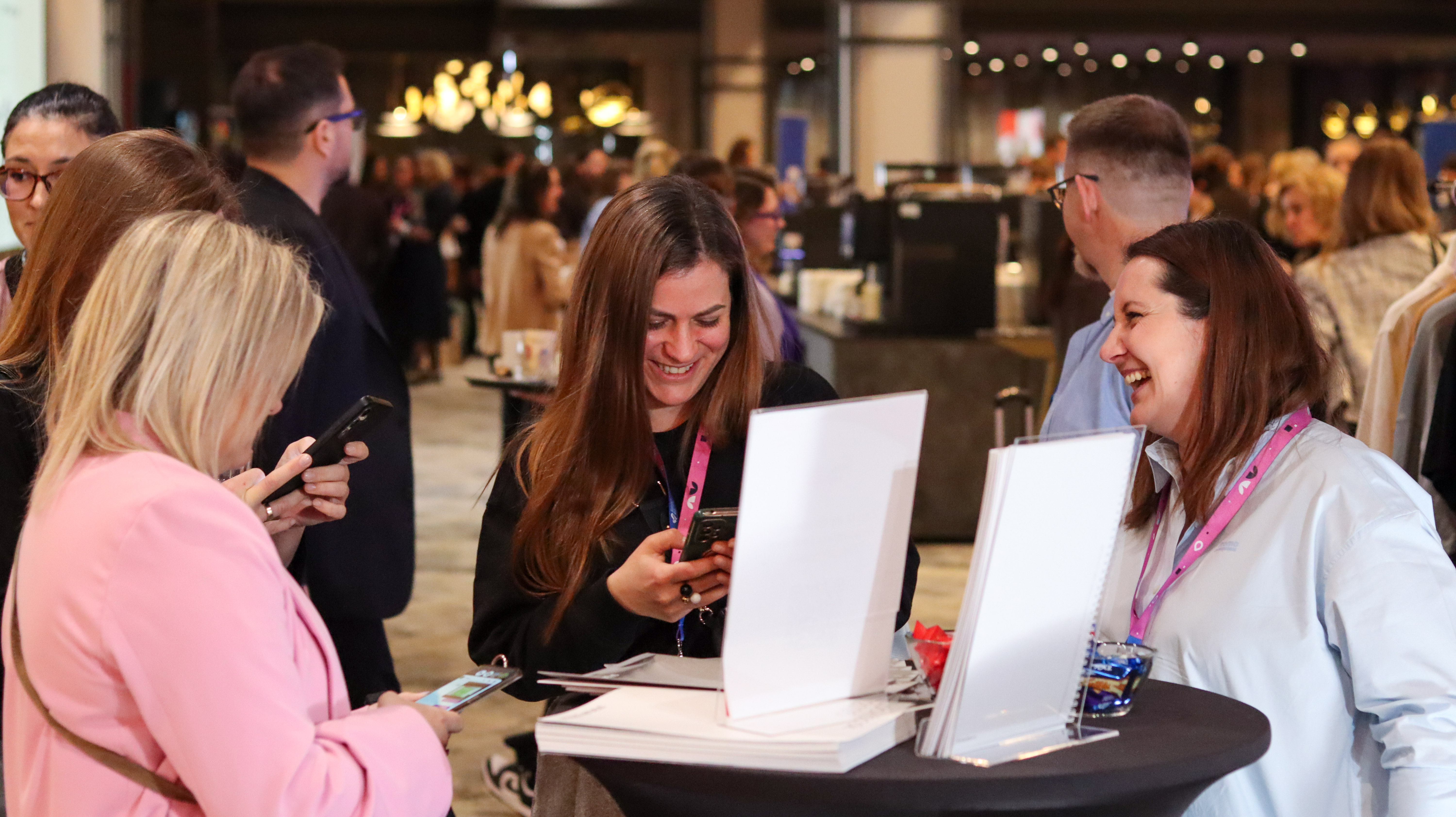 Kobieta w błękitnej koszuli firmowej i jeansach uśmiecha się szeroko, rozmawiając z inną kobietą przy czarnym stoliku koktajlowym. Na stole widać ulotki i ekspozycję. Atmosfera networkingowa.