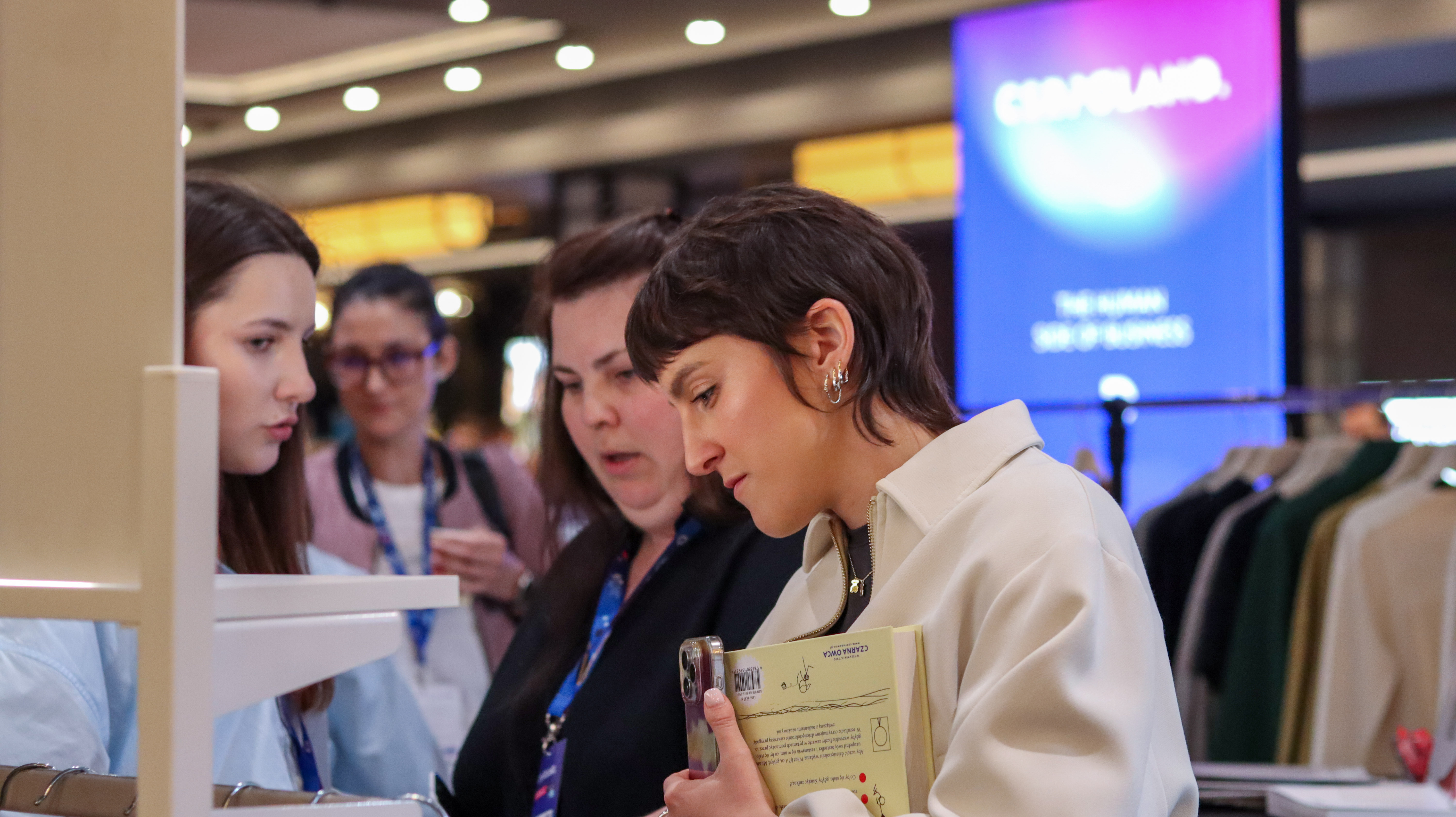 Kobieta o kr&oacute;tkich włosach w jasnej kurtce, trzymająca w dłoni telefon i książkę, uważnie ogląda ekspozycję na stoisku. Obok niej stoją dwie inne kobiety. W tle widać niebieski świecący baner z napisem &bdquo;CSR POLAND&rdquo; oraz &bdquo;THE HUMAN SIDE OF BUSINESS&rdquo;.