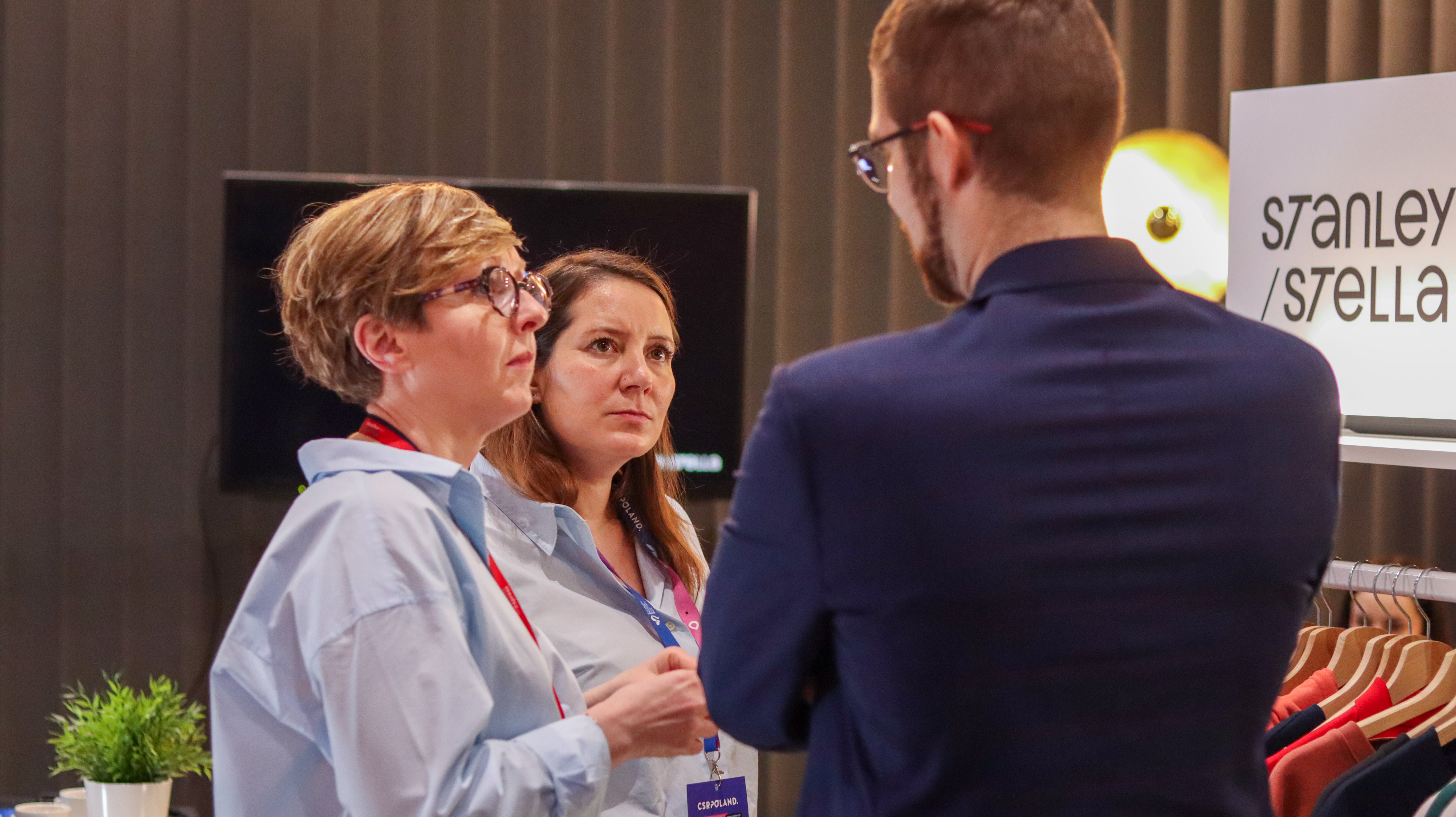 Kobieta w okularach i błękitnej koszuli rozmawia z uczestnikami wydarzenia. W tle widoczny baner Promo Express oraz wieszak z ekologiczną odzieżą marki Stanley/Stella.