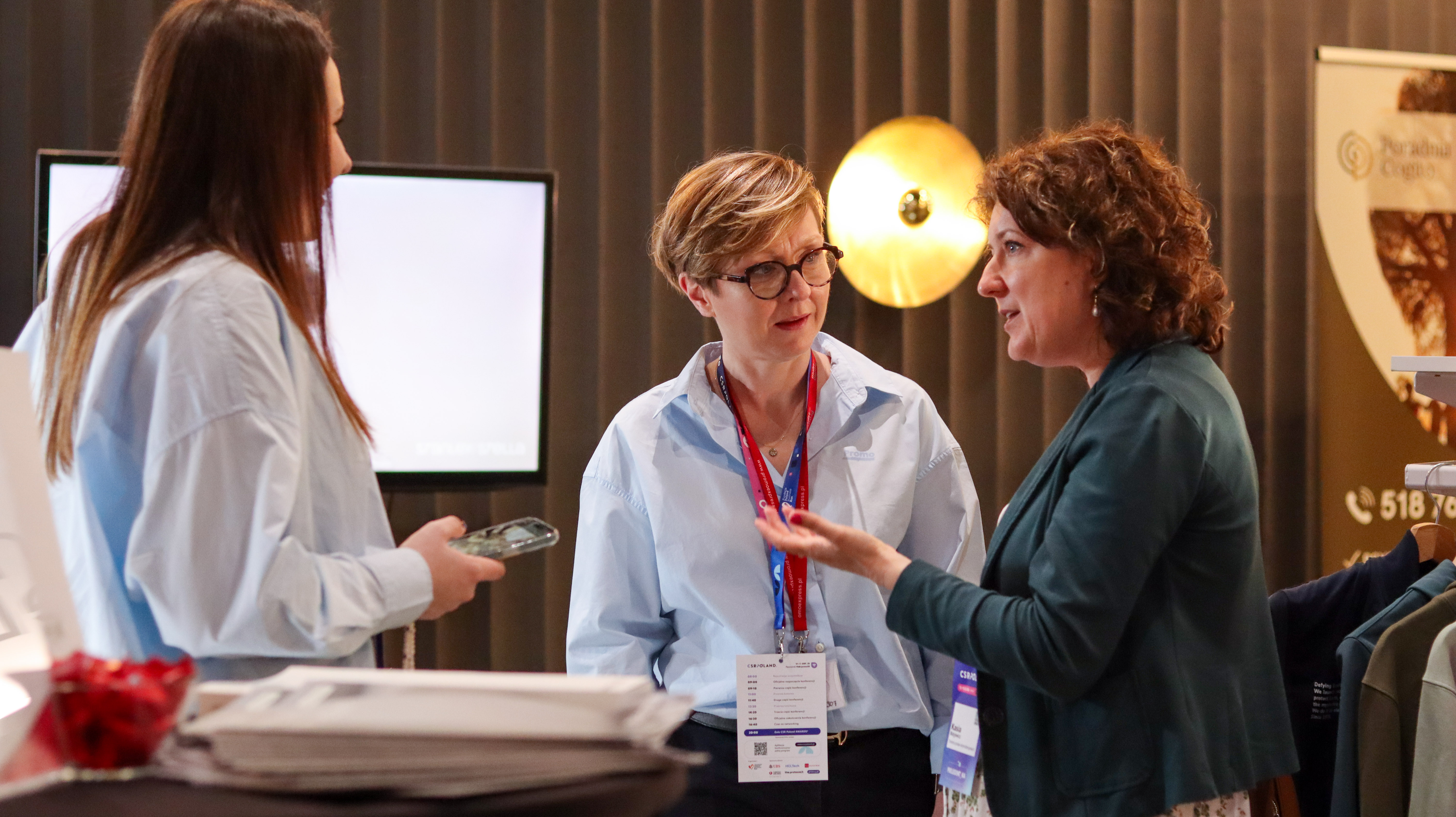 Trzy kobiety rozmawiają w kuluarach konferencji. Jedna z nich ma na szyi smycz z identyfikatorem. W tle nowoczesne wnętrze z monitorem i złotą lampą.