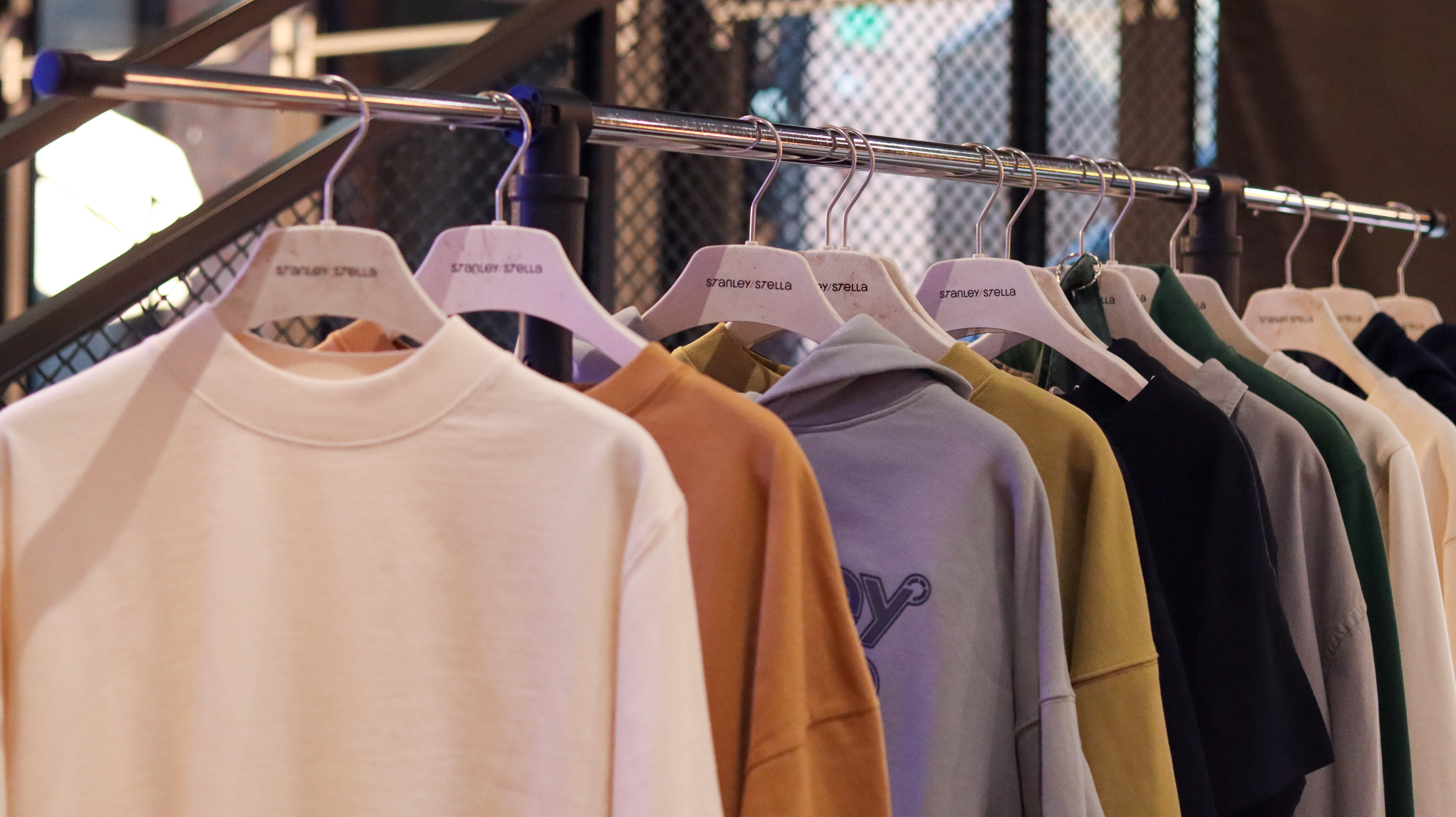 Zbliżenie na markowe bluzy i koszulki zawieszone na białych wieszakach z logo Stanley/Stella. Odzież w odcieniach beżu, pomarańczu i szarości.