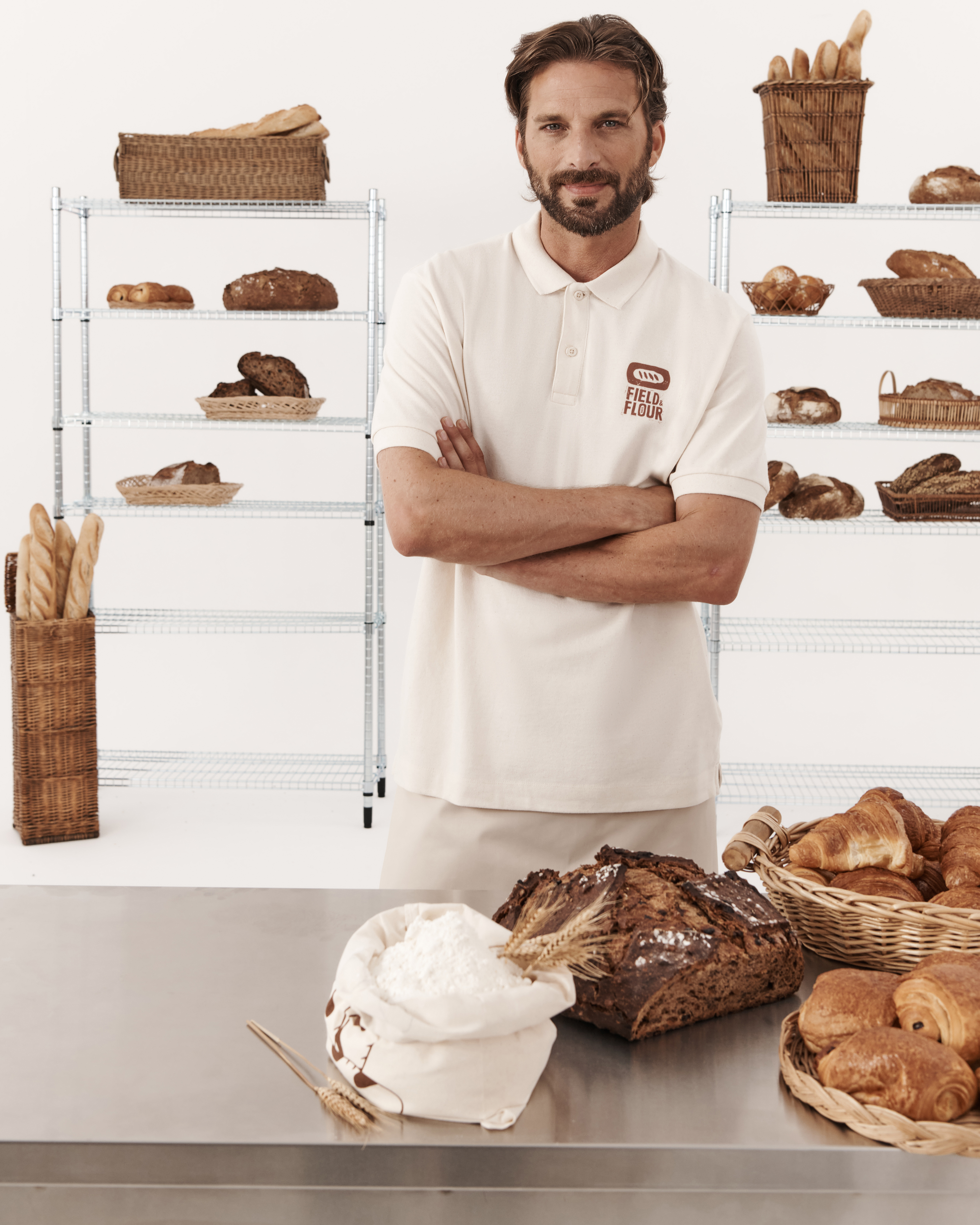 Mężczyzna z brodą w kremowej koszulce polo z logo &bdquo;FIELD & FLOUR&rdquo; stoi z założonymi rękami w piekarni na tle regał&oacute;w z pieczywem.