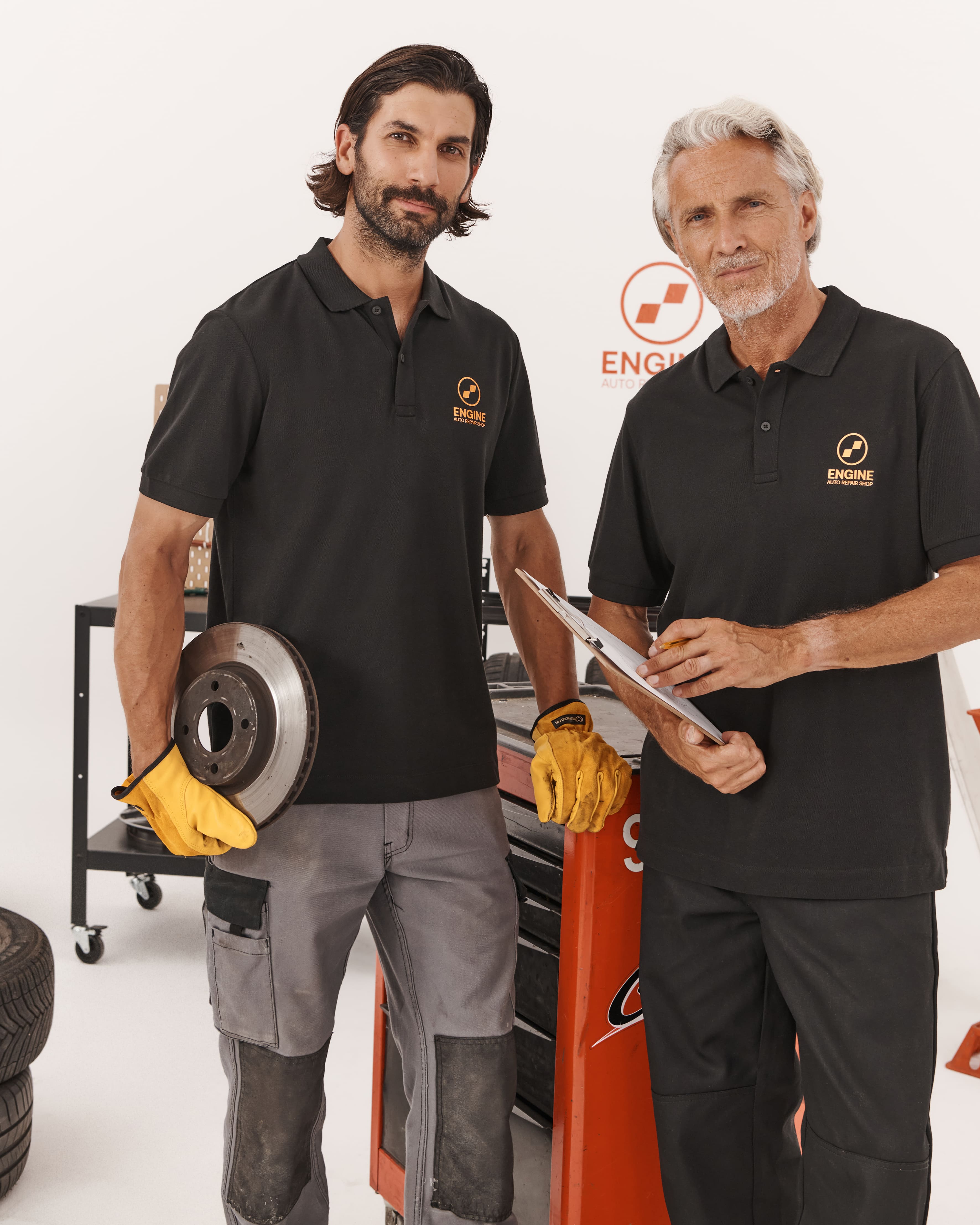 Dwaj mężczyźni w czarnych koszulkach polo z logo &bdquo;ENGINE&rdquo; w warsztacie; jeden trzyma tarczę hamulcową, drugi podkładkę z dokumentami.