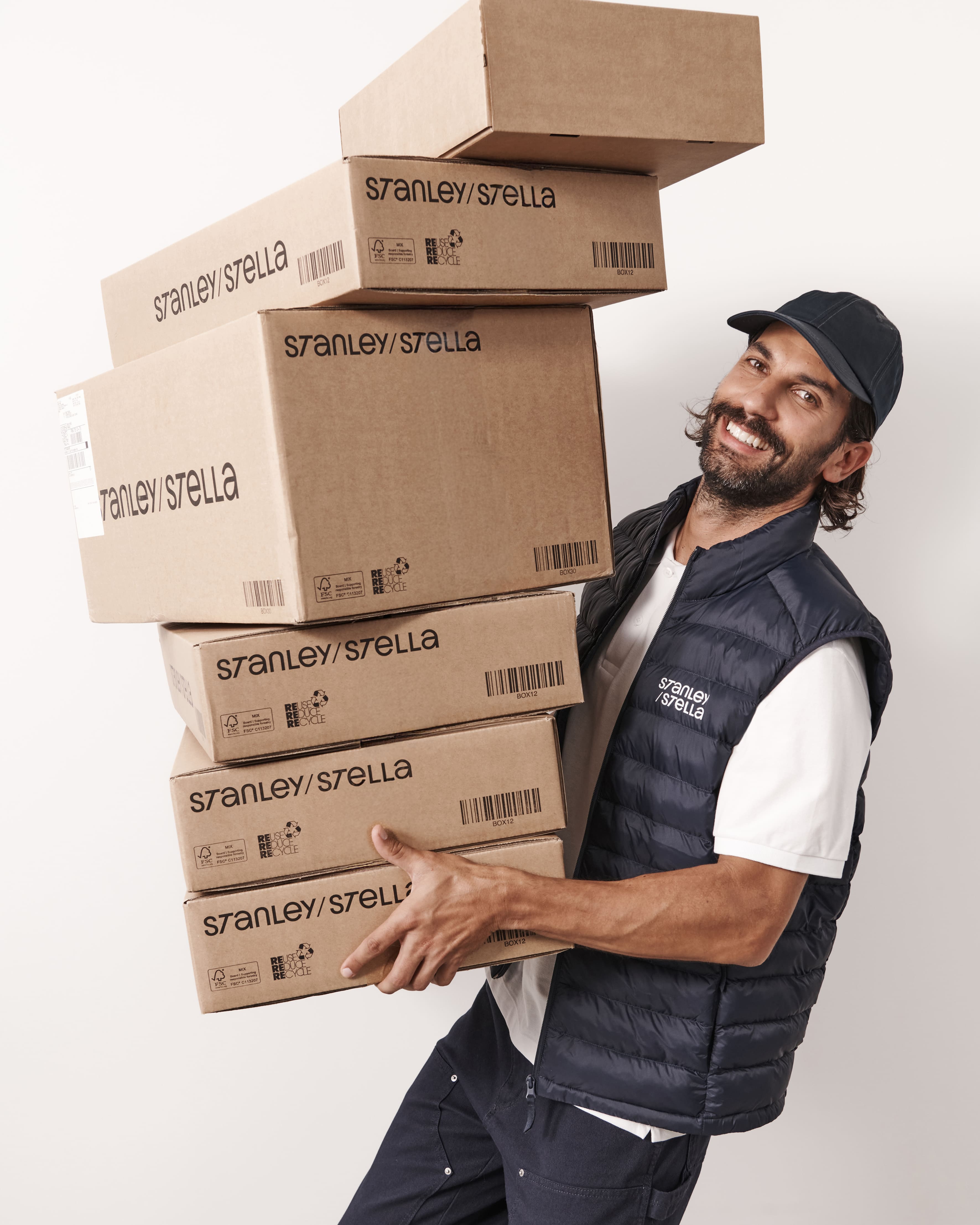 Uśmiechnięty mężczyzna w granatowej kamizelce i czapce z daszkiem niesie wysoki stos kartonowych pudeł z logo &bdquo;Stanley/Stella&rdquo;.