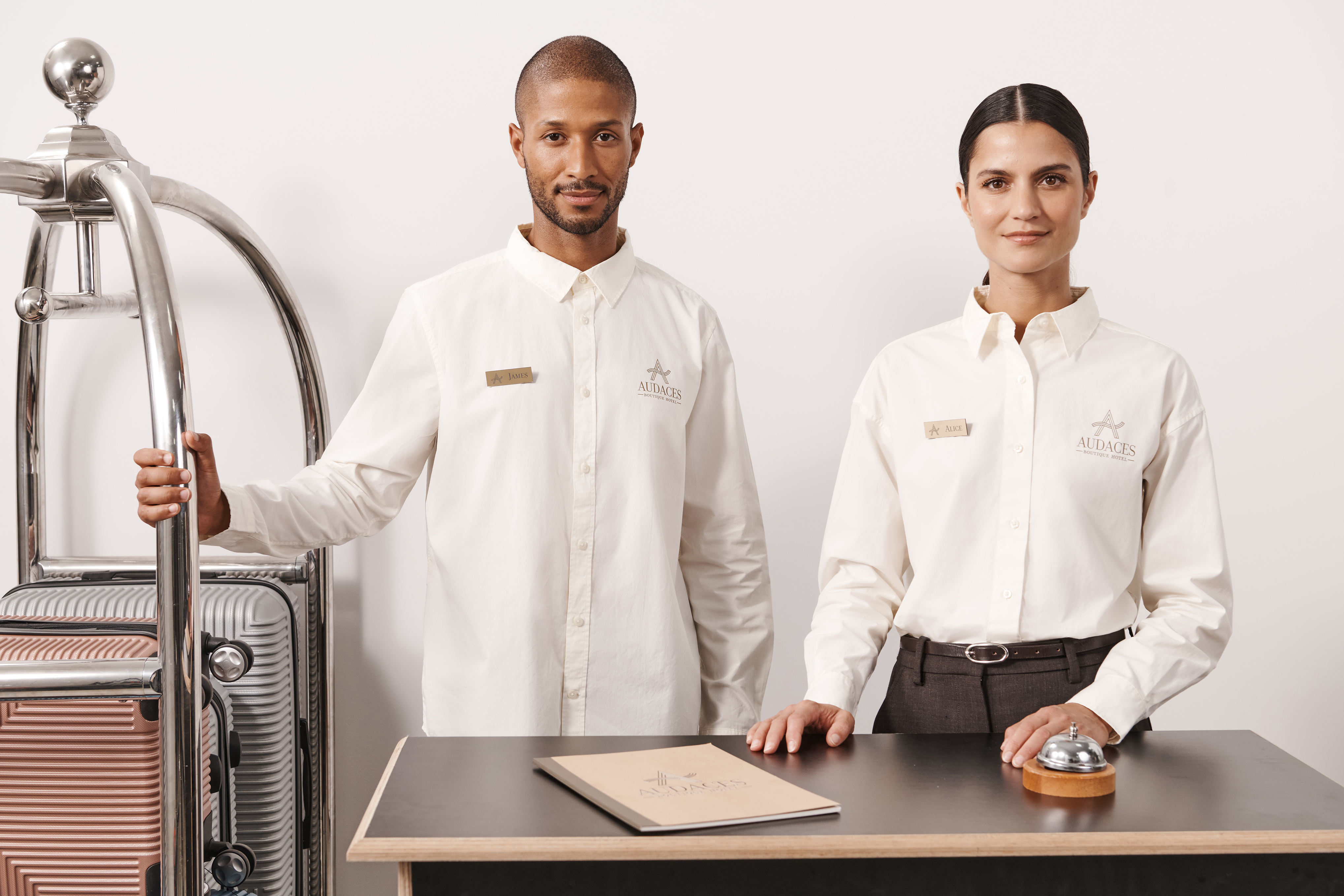 Kobieta i mężczyzna w eleganckich, białych koszulach z logo &bdquo;AUDACES&rdquo; stoją za hotelową ladą recepcyjną obok w&oacute;zka na bagaż.