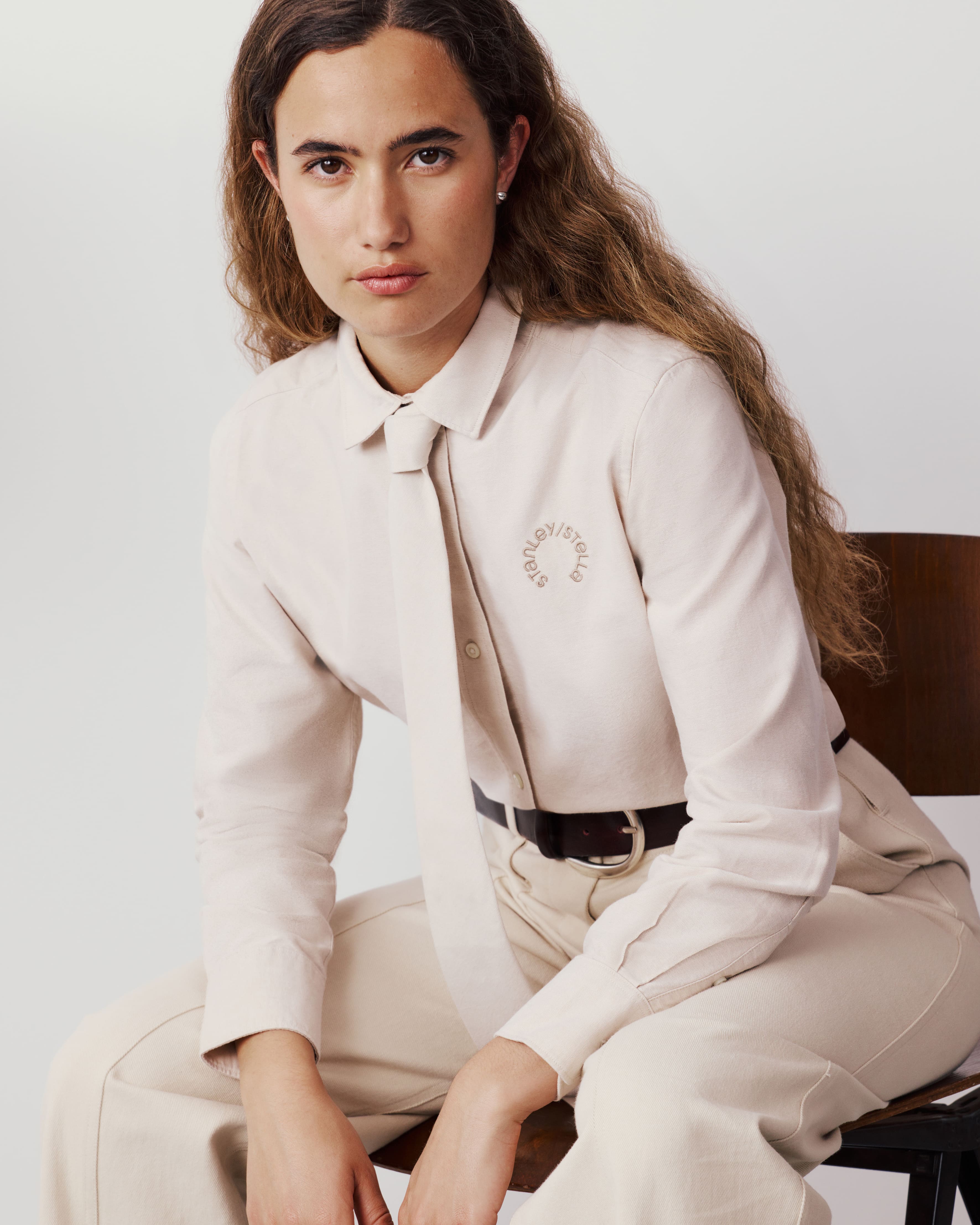 Kobieta siedząca na krześle, ubrana w jasną koszulę z krawatem w tym samym kolorze i subtelnym haftem na piersi.