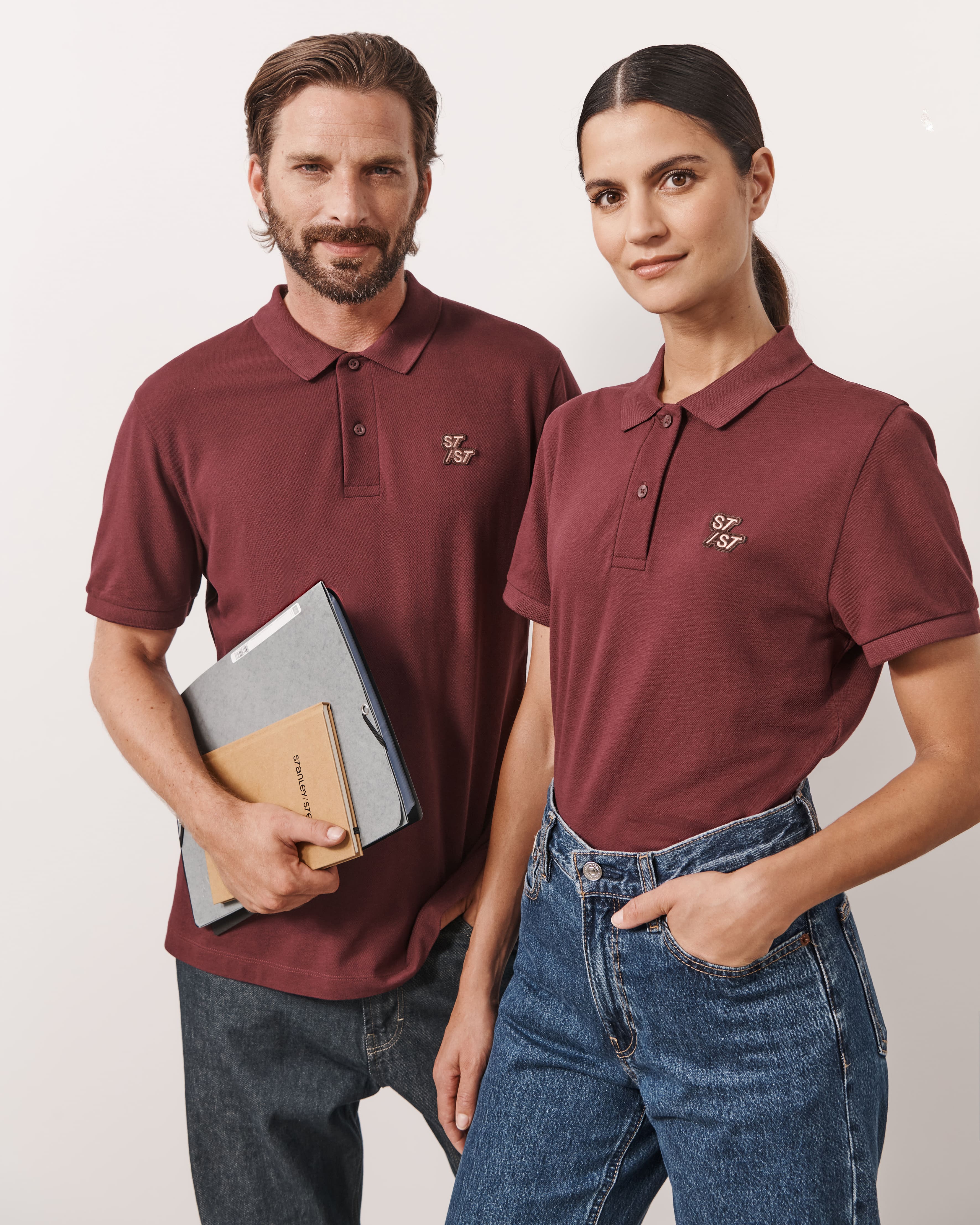 Kobieta i mężczyzna w dopasowanych, bordowych koszulkach polo z małym logo na piersi, trzymający foldery i notatniki.
