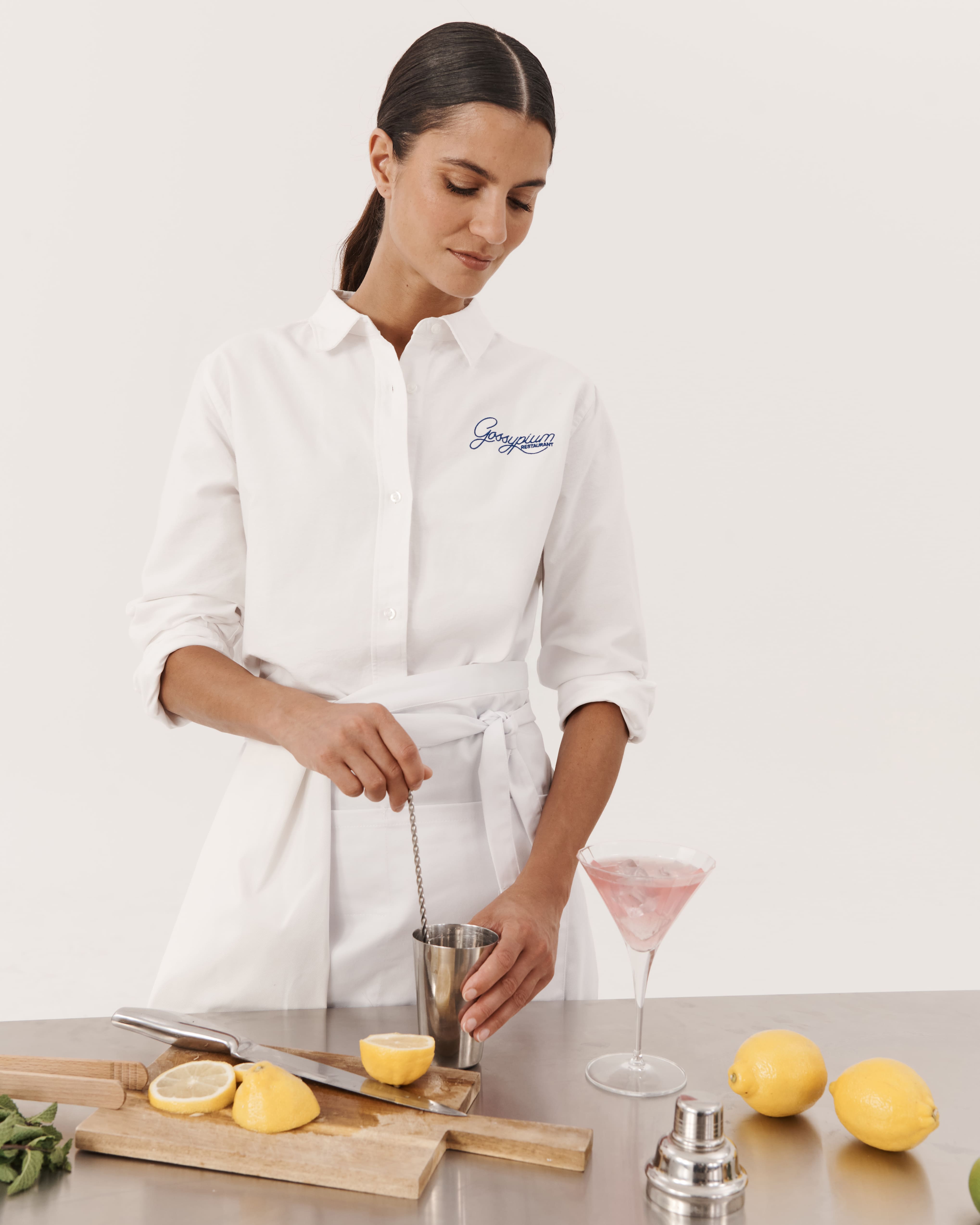 Kobieta w białej koszuli Oxford z logo &bdquo;Gossypium&rdquo; i białym fartuchu, przygotowująca koktajl przy stalowym blacie z cytrynami i akcesoriami barmańskimi.