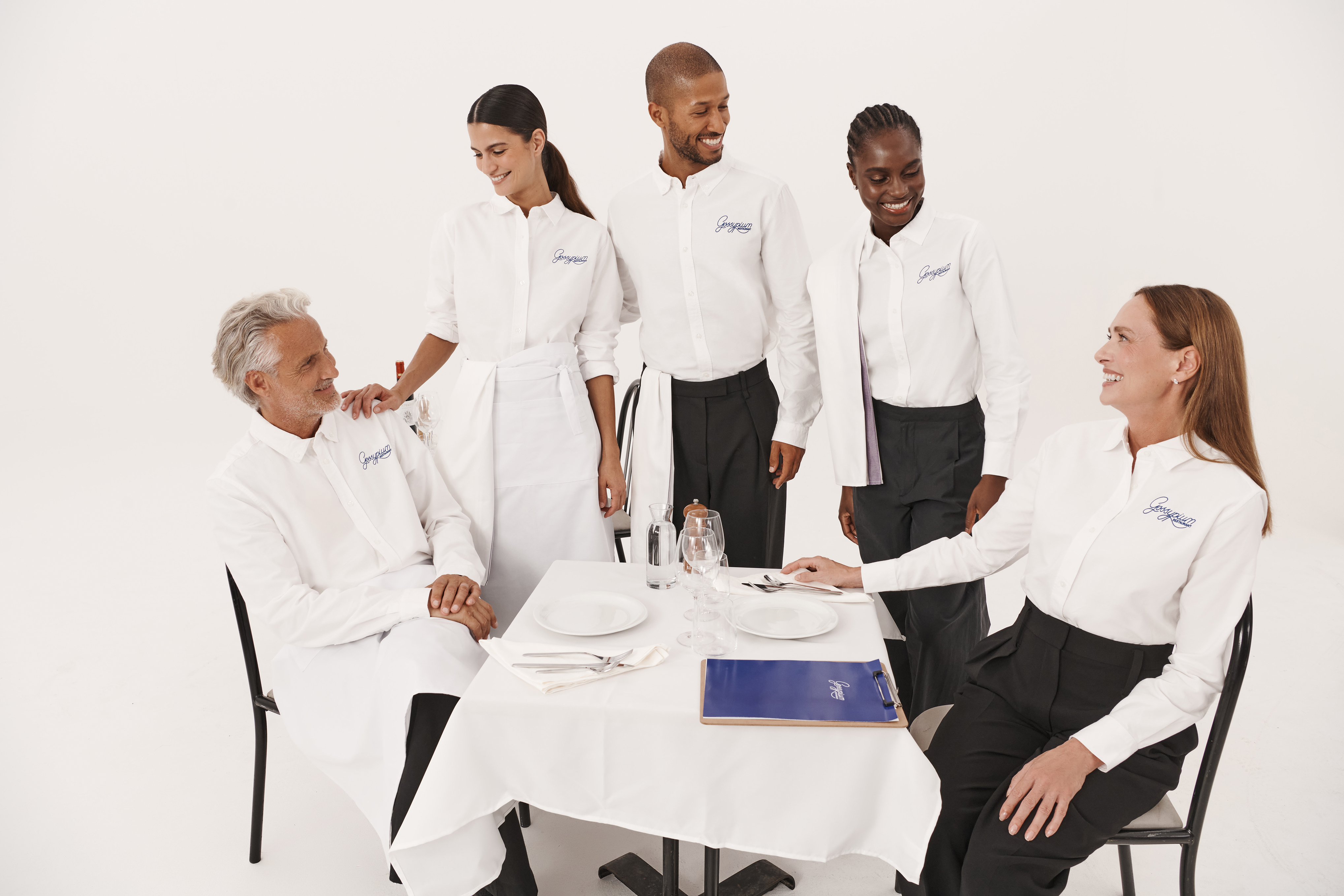 Grupa os&oacute;b w białych koszulach z niebieskim logo, stylizowanych na obsługę restauracji, zgromadzona wok&oacute;ł stolika w białym studio.